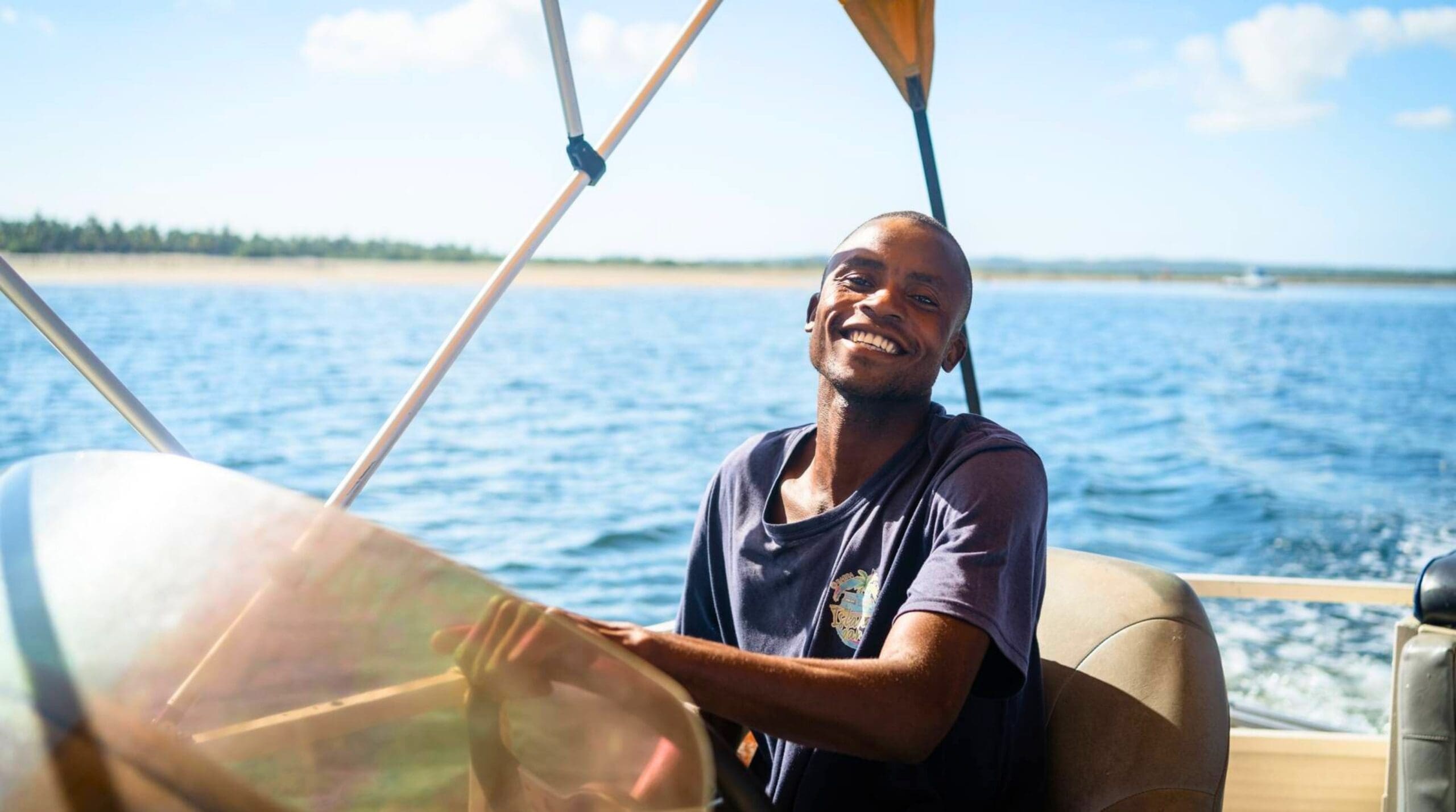 Een lachende man in een marine t-shirt bestuurt een boot op een zonnige dag, met blauw water en een verre kustlijn zichtbaar op de achtergrond.