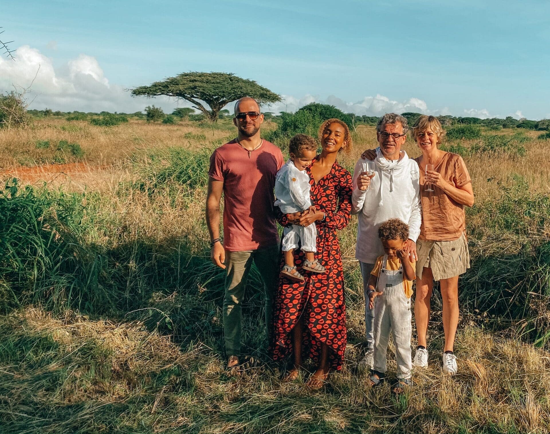 familie met jonge kinderen poseert in grasvlakte met wijnglazen in de hand tijdens safari in Kenia