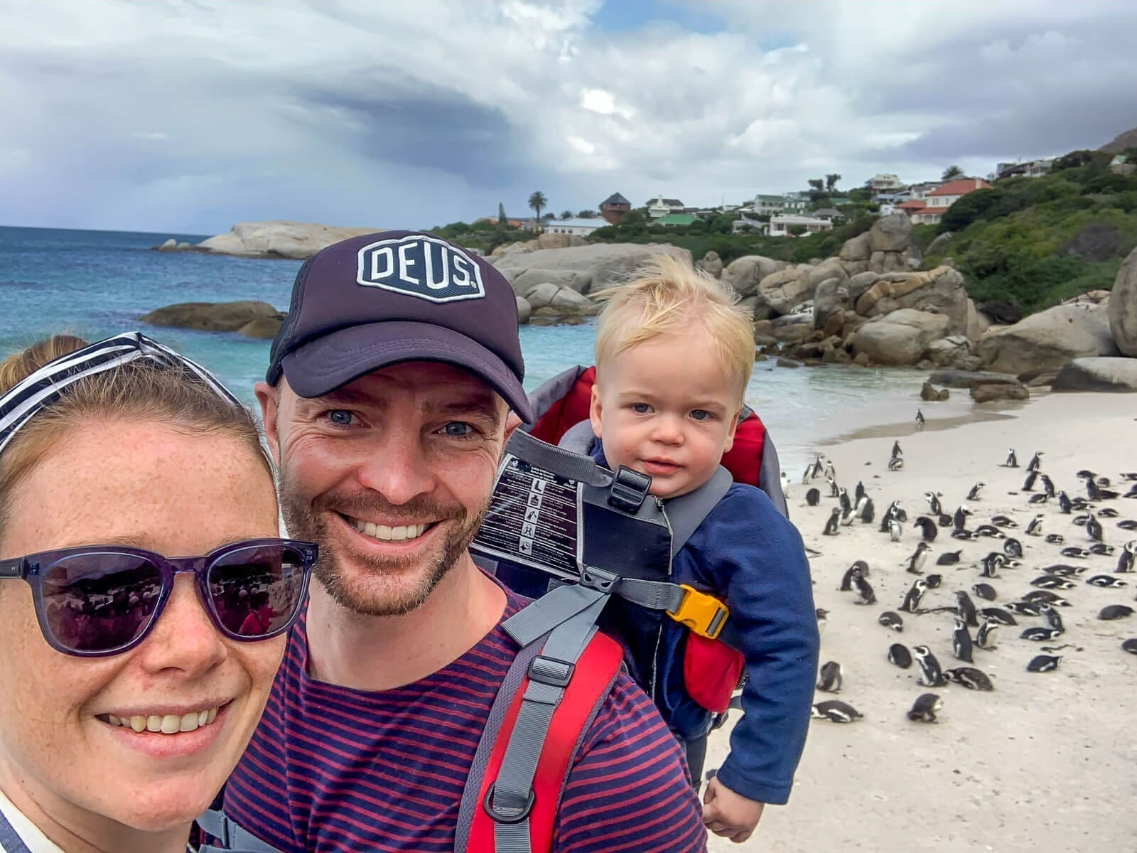 Een lachend stel neemt een selfie met hun peuter in een draagzak op een zandstrand. Tientallen pinguïns zitten op het zand achter hen, met rotsachtige kliffen, oceaangolven en huizen op de achtergrond.
