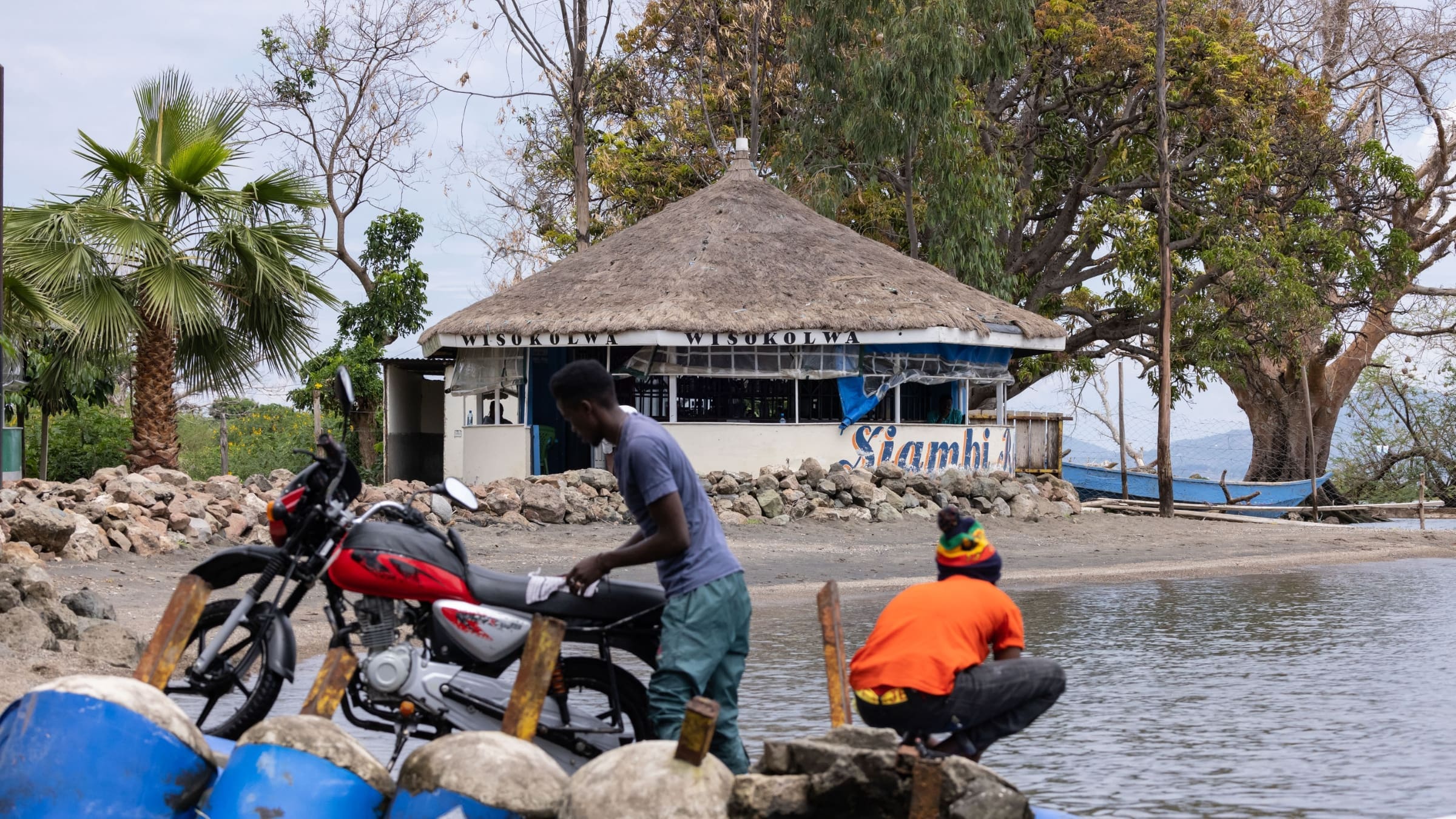 Twee mannen aan de oever bij een motorfiets met op de achtergrond een traditioneel rond huis en Lake Naivasha