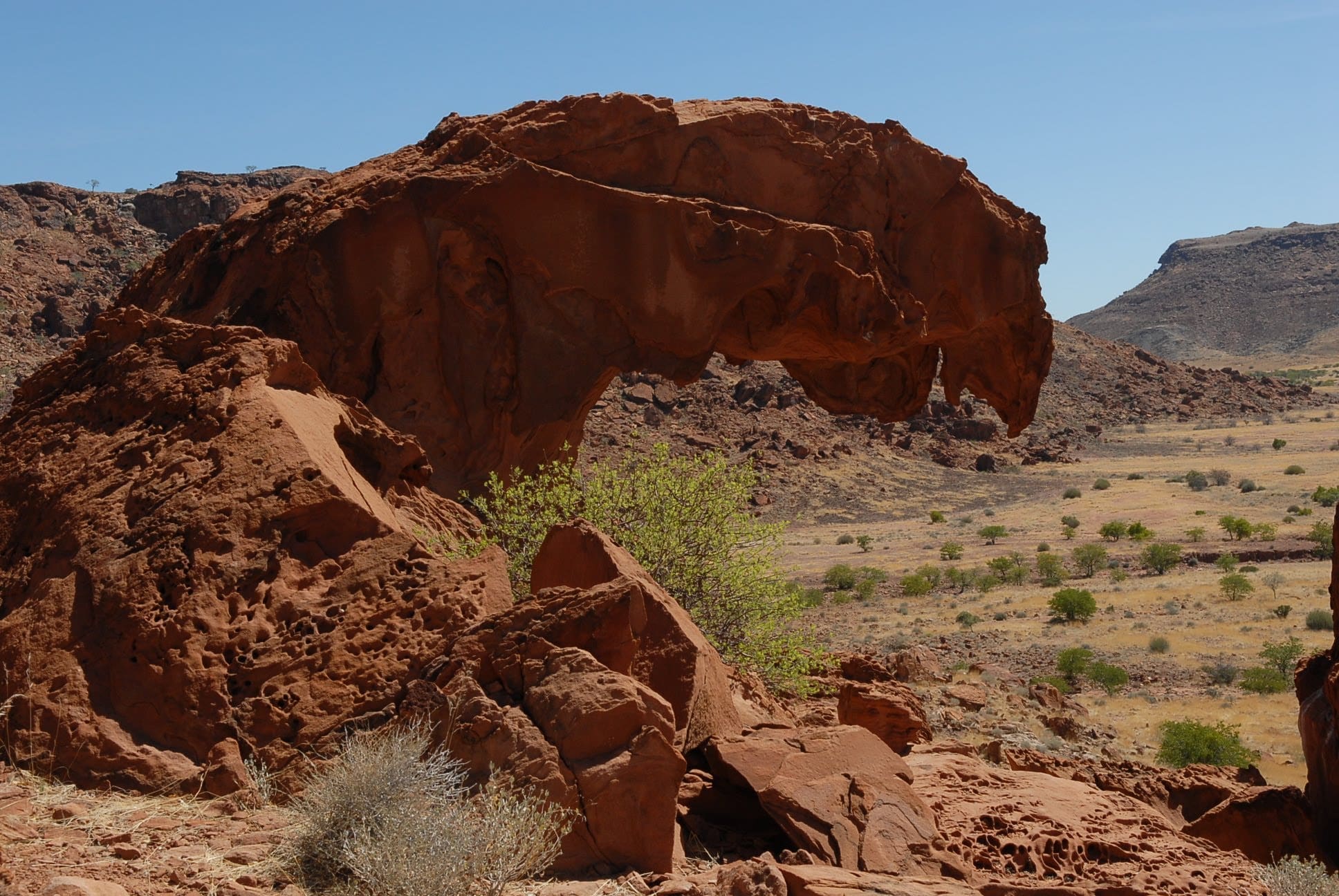 A large, reddish-brown rock formation with a curved, arch-like shape rises above a dry, grassy landscape with sparse green shrubs under a clear blue sky.