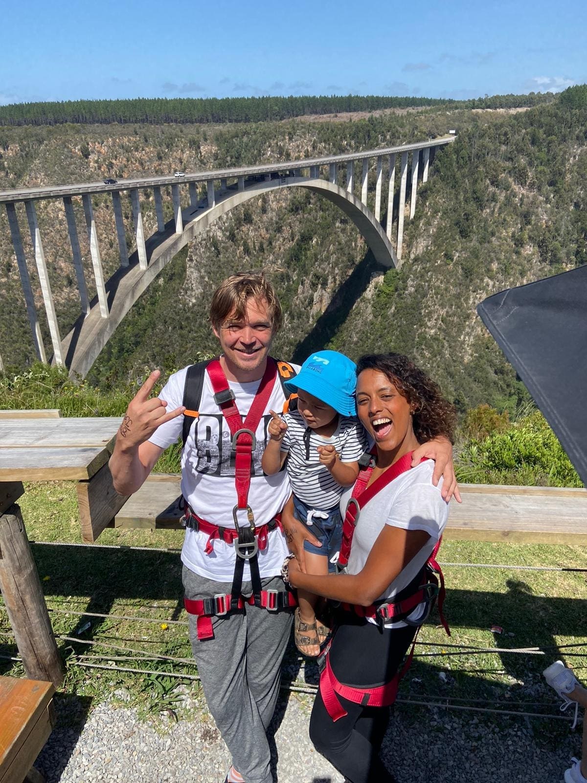 Drie mensen met een harnas aan glimlachen voor een foto voor een schilderachtige, hoge boogbrug over een diepe kloof met groen. De man geeft een vredesteken, de vrouw houdt een kind met een blauwe hoed vast en het is een zonnige dag.