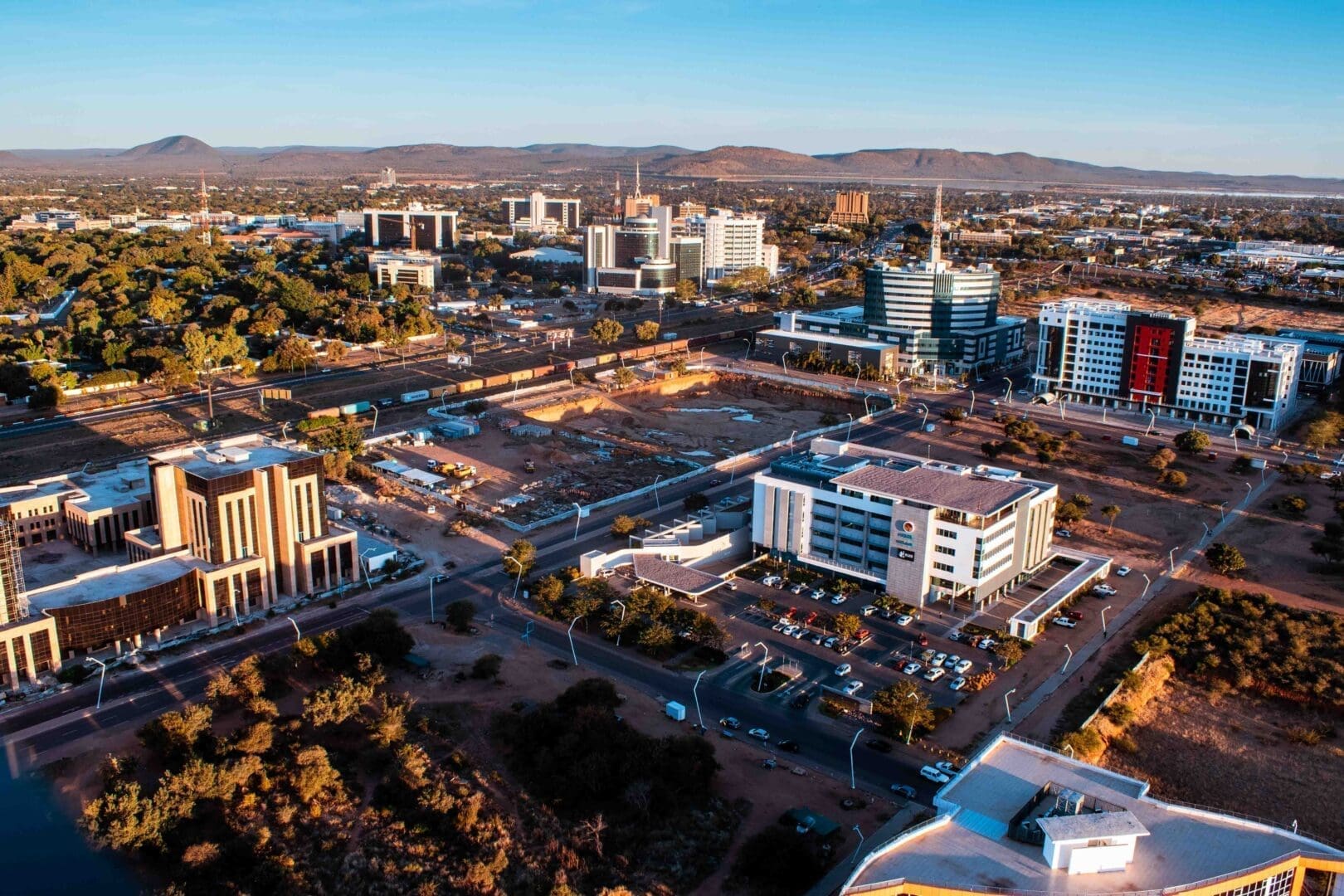 Aerial view of a modern city in Botswana, Africa, with office buildings, roads, parking lots, and scattered trees under a clear blue sky—perfect for those travelling and seeking scenic urban landscapes with distant hills.