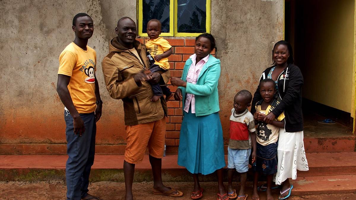 Zes mensen, waaronder twee mannen, twee vrouwen en twee jonge kinderen, staan glimlachend voor een huis met een gele deur. Deze vrolijke reizigers lijken klaar voor hun volgende avontuur-misschien zelfs een reis naar Oeganda!.