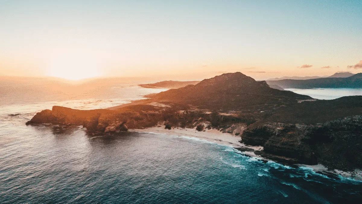 Luchtfoto van de ruige kustlijn van Zuid-Afrika bij zonsondergang, met golven die op rotsachtige kliffen slaan en een bergachtig landschap - een droombestemming voor reizigers die van avontuurlijk reizen houden.
