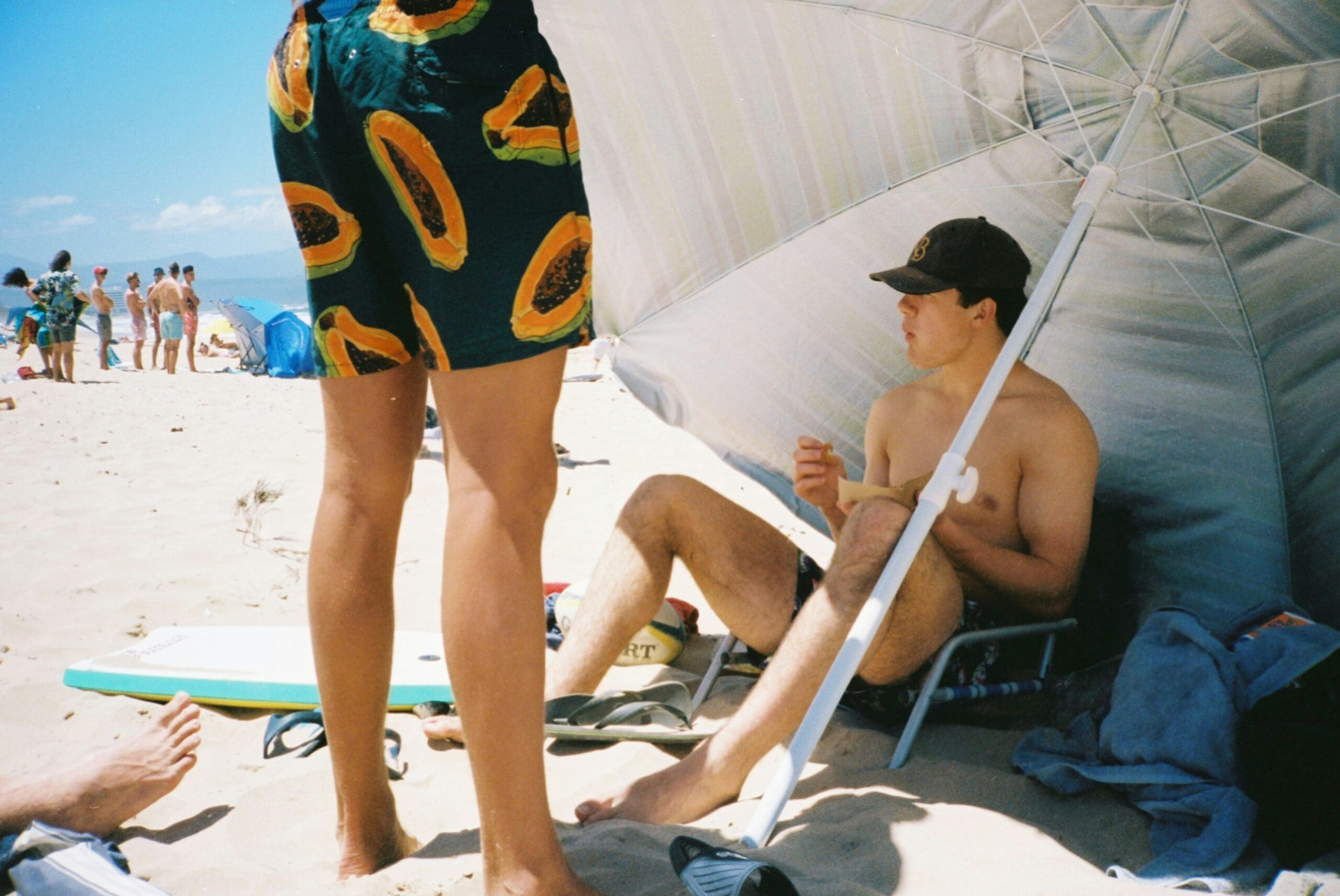 Een shirtloze persoon zit onder een grote parasol op het zand, terwijl een andere persoon in een korte broek met fruitmotief vlakbij staat. Andere strandgangers en surfplanken zijn zichtbaar op de achtergrond op een zonnige dag.