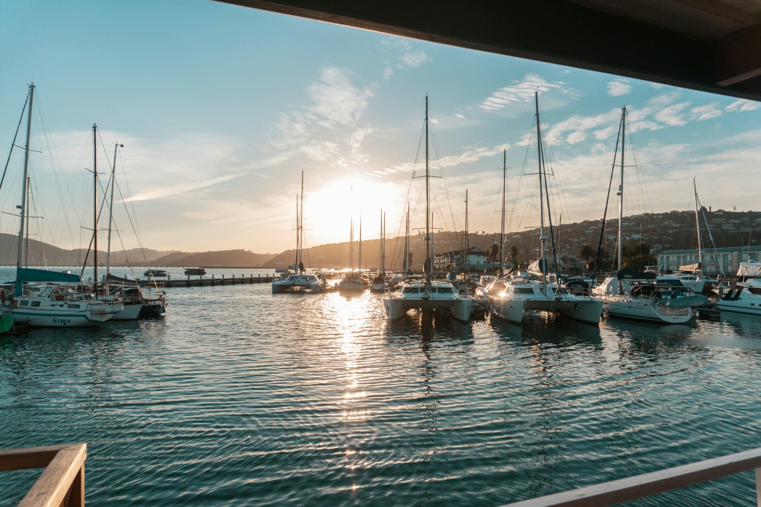 Zeilboten aangemeerd in een jachthaven tijdens zonsondergang, met zonlicht dat reflecteert op het kalme water en de heuvels op de achtergrond. Het beeld wordt omlijst door een schaduwrijk bouwwerk op de voorgrond.