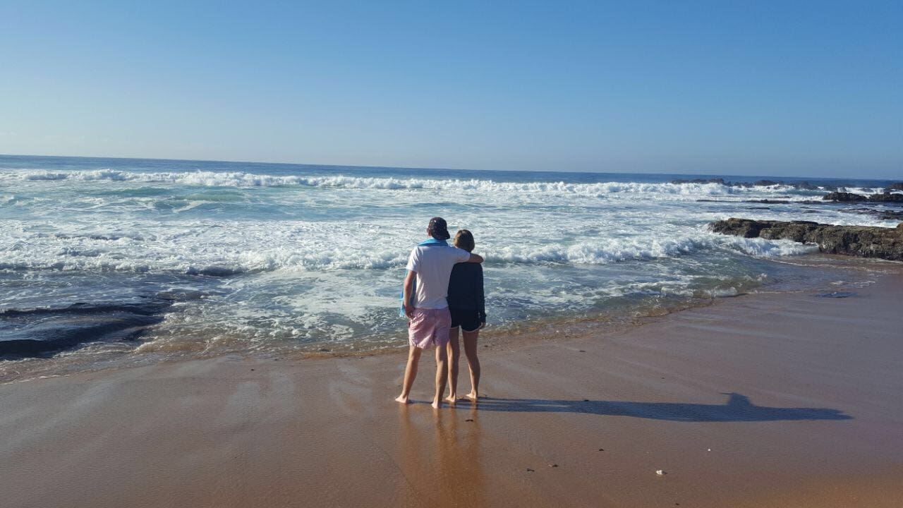 Een stel staat op blote voeten op een zandstrand, uitkijkend op de oceaan met golven die op de kust slaan onder een strakblauwe hemel. Ze omhelzen elkaar en creëren een vredig, intiem tafereel.