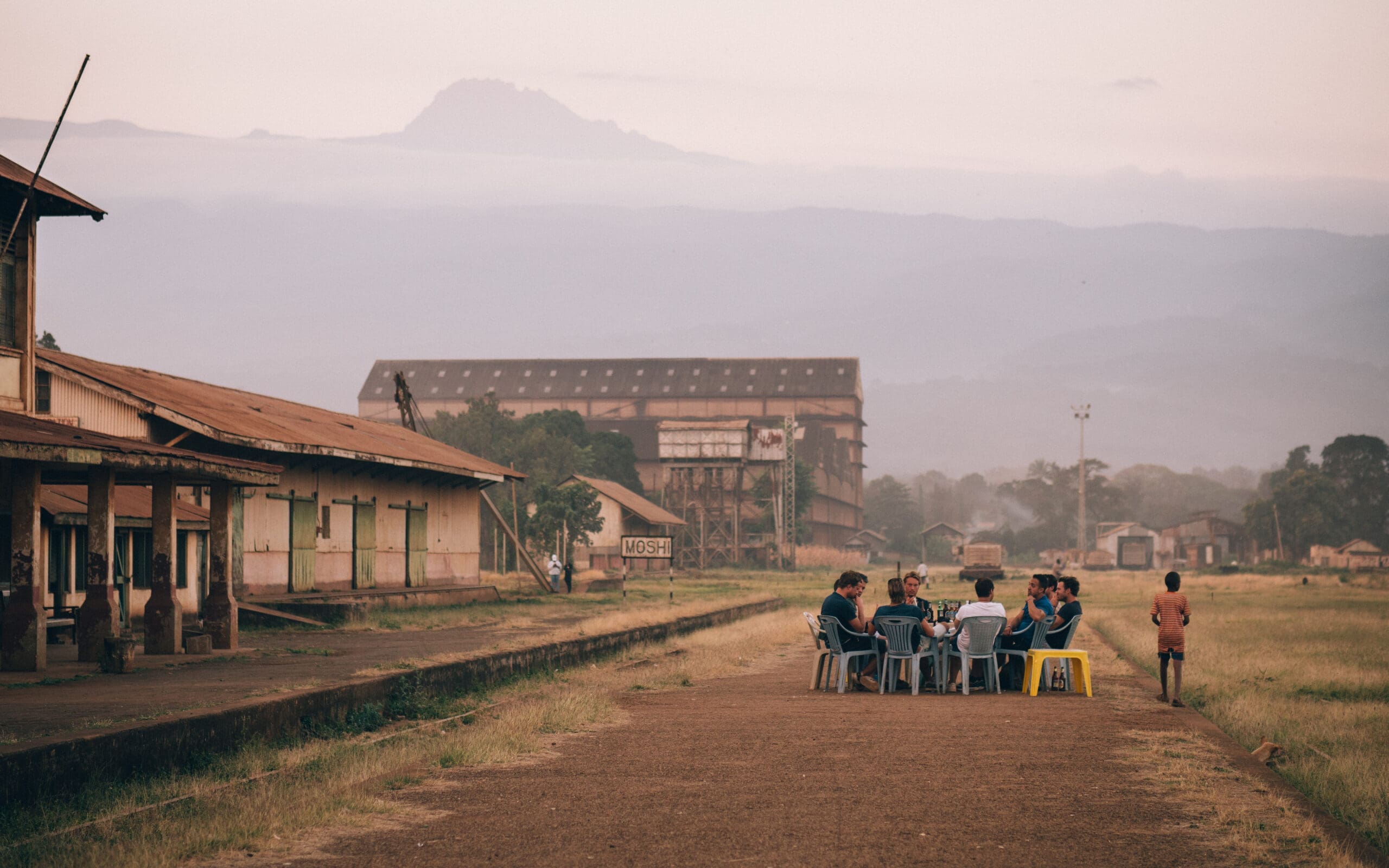 Een groep mensen zit in plastic stoelen rond een tafel buiten bij oude gebouwen op het platteland van Tanzania, met bergen op de achtergrond en een kind dat vlakbij op een zandpad staat. Het kalme Afrika tafereel baadt in het licht van de zonsondergang, perfect voor wie van reizen houdt.
