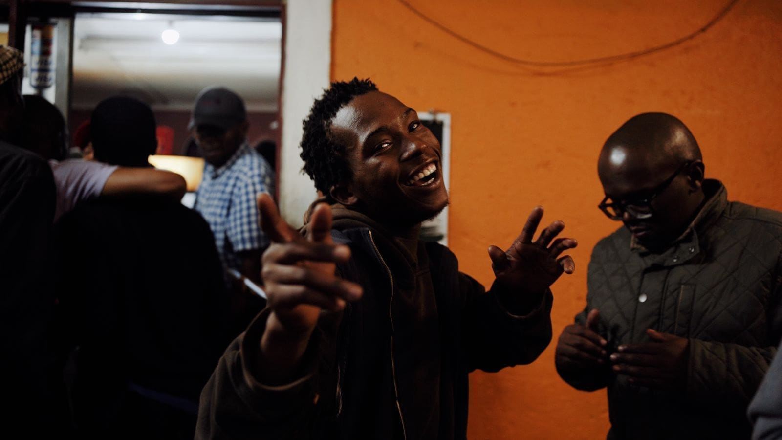 een jongeman lacht en wijst naar de camera terwijl hij zich onder een groep mensen bevindt tijdens een avondmoment in eswatini