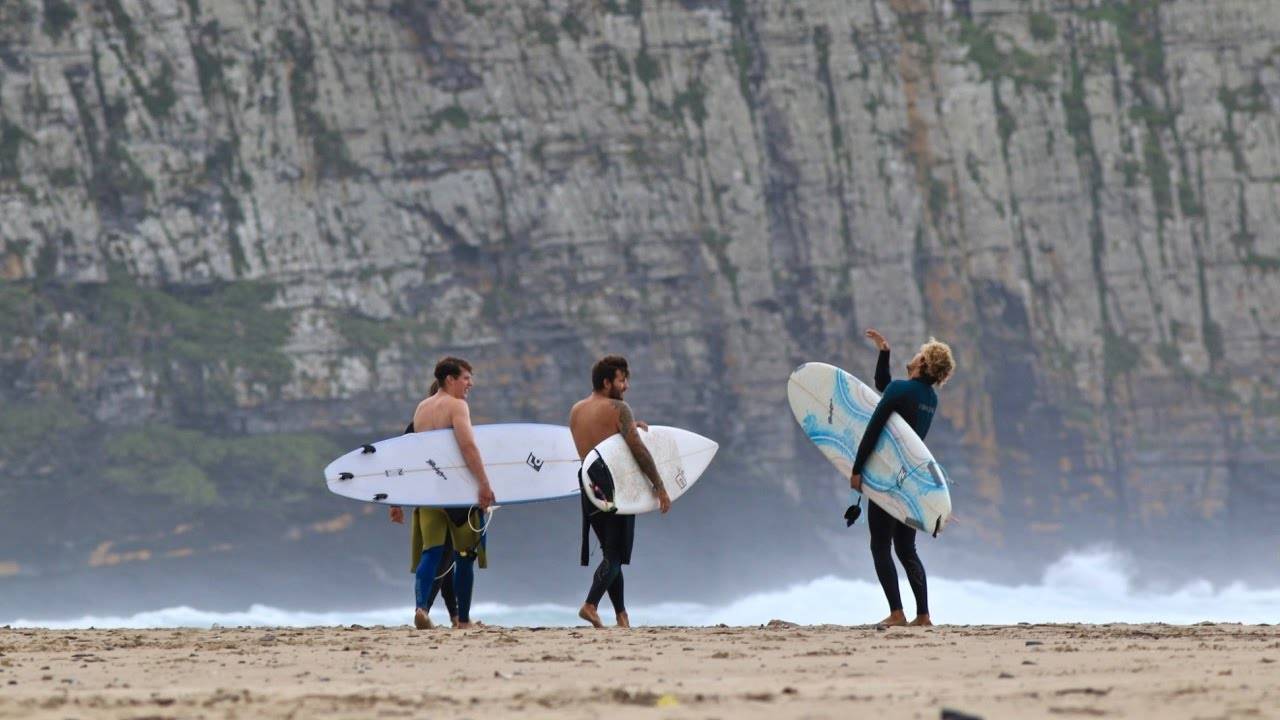 Drie reizigers in wetsuits staan op een zandstrand met surfplanken in de hand en kletsen terwijl de oceaangolven tegen een grote rotswand in Zuid Afrika beuken.