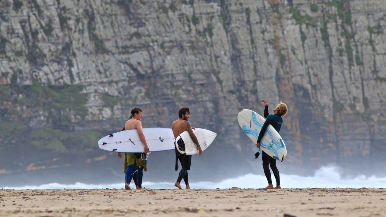 Drie surfers in wetsuit staan op een zanderig Zuid-Afrikaans strand met surfplanken en kletsen terwijl hoge rotswanden en golven de perfecte achtergrond vormen voor een onvergetelijk reisavontuur.