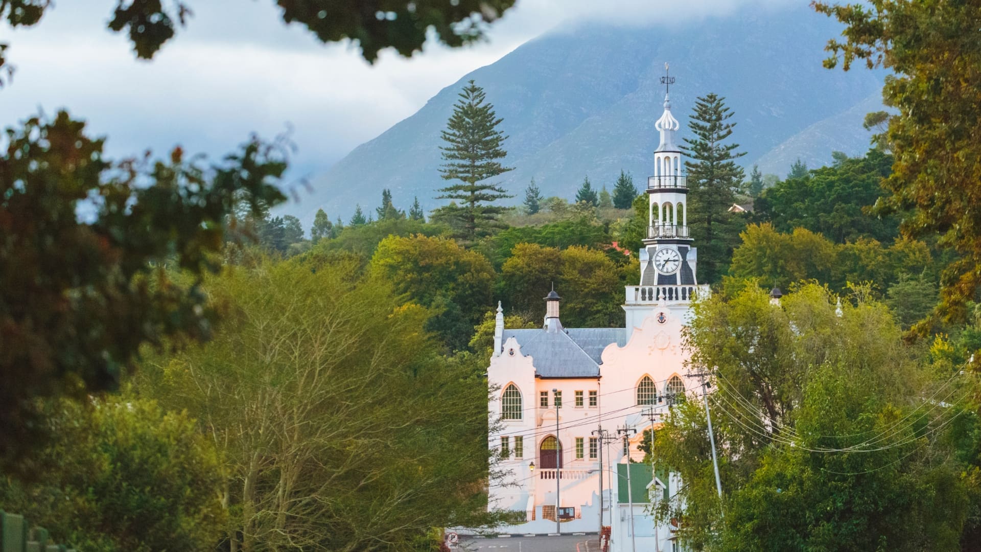 Een witte kerk met een hoge klokkentoren staat tussen groene bomen, met bergen op de achtergrond onder een bewolkte hemel.