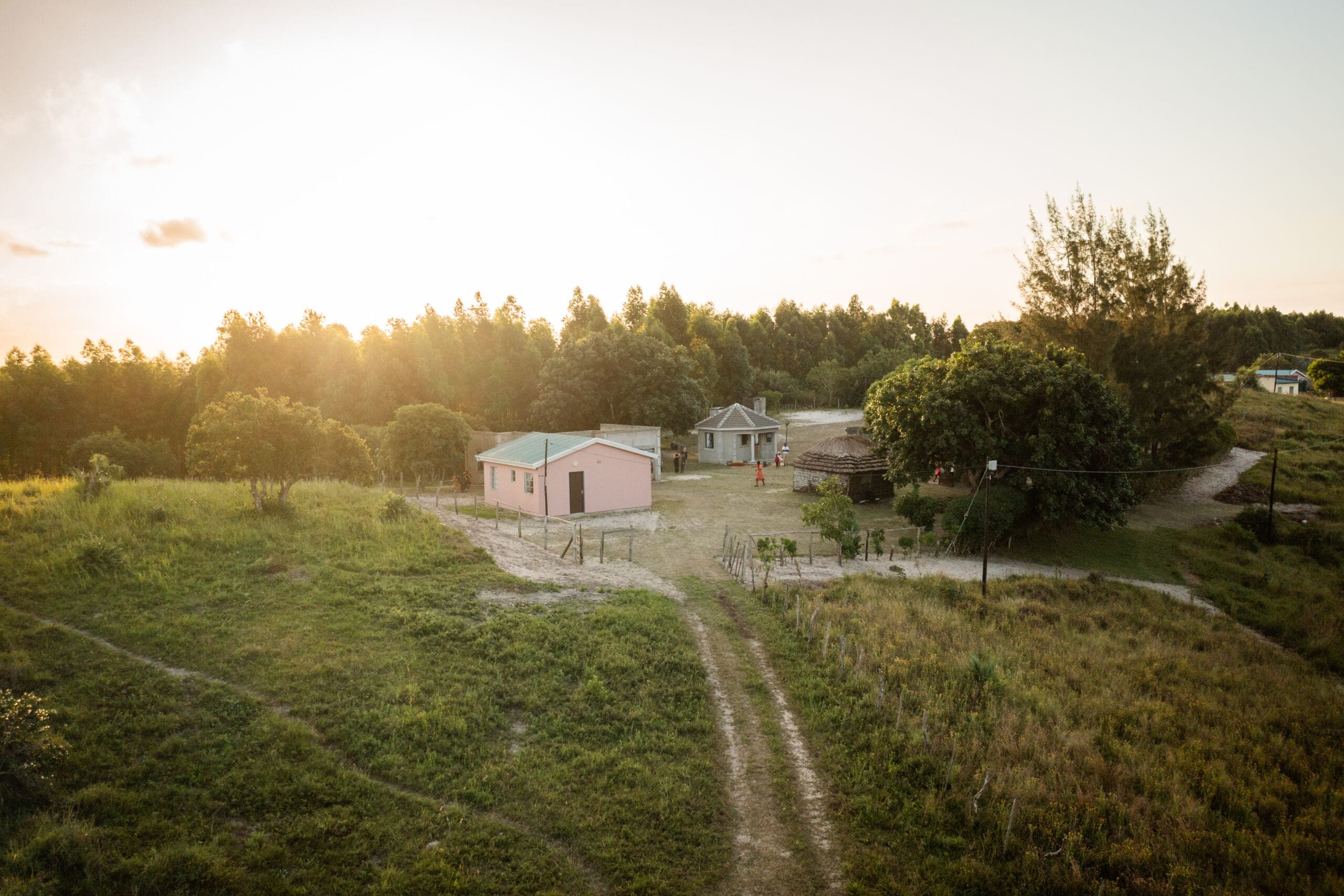 Eine ländliche Szene bei Sonnenuntergang mit einem rosafarbenen Haus und einem kleineren grauen Gebäude, umgeben von Gras, Bäumen und unbefestigten Wegen, vor dem Hintergrund eines dichten Waldes.