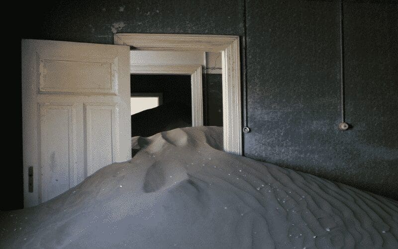 Een kamer gevuld met zandduinen die tot halverwege een witte deur reiken, met meer zand zichtbaar in de volgende kamer. De muren zijn donker en op de muren zijn oude lichtschakelaars en blootliggende draden te zien.