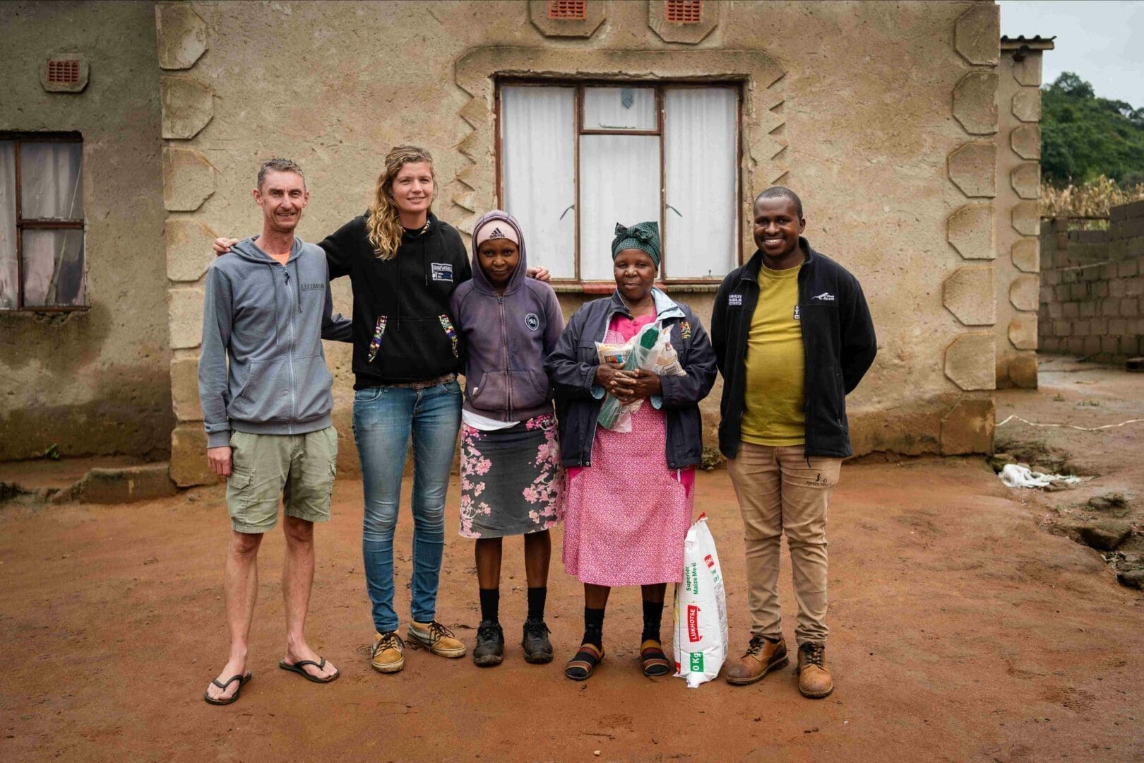 een groep van vijf volwassenen staat samen voor een traditionele woning in eswatini waarbij ze lachen en verbondenheid uitstralen tijdens een lokale gemeenschapsactiviteit