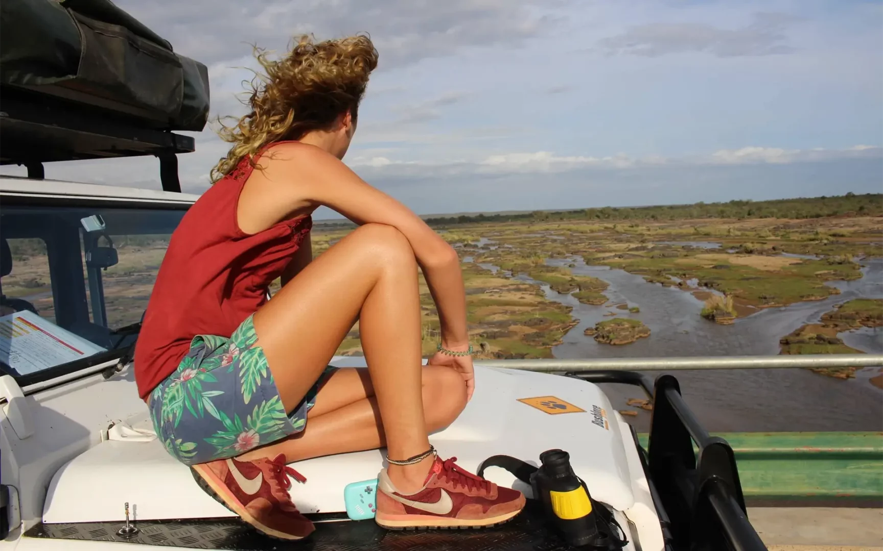 Een persoon met krullend haar in een rood tanktopje en een gebloemde korte broek zit op de motorkap van een voertuig en kijkt uit over een schilderachtig rivierlandschap - een uitnodigend uitzicht voor reizigers die op zoek zijn naar avontuur in Afrika onder een gedeeltelijk bewolkte hemel.