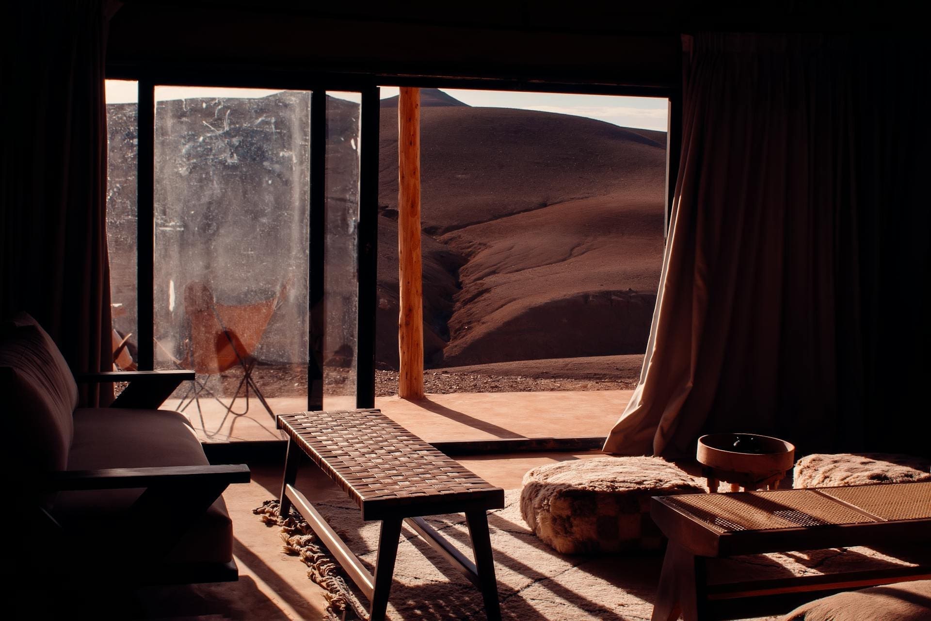 Een gezellige kamer met houten meubels en poefs kijkt uit op grote glazen deuren die openen naar een patio en zonovergoten, dorre woestijnheuvels onthullen - een uitnodigend toevluchtsoord voor Afrika reizigers. Warme, zachte verlichting vult het interieur.