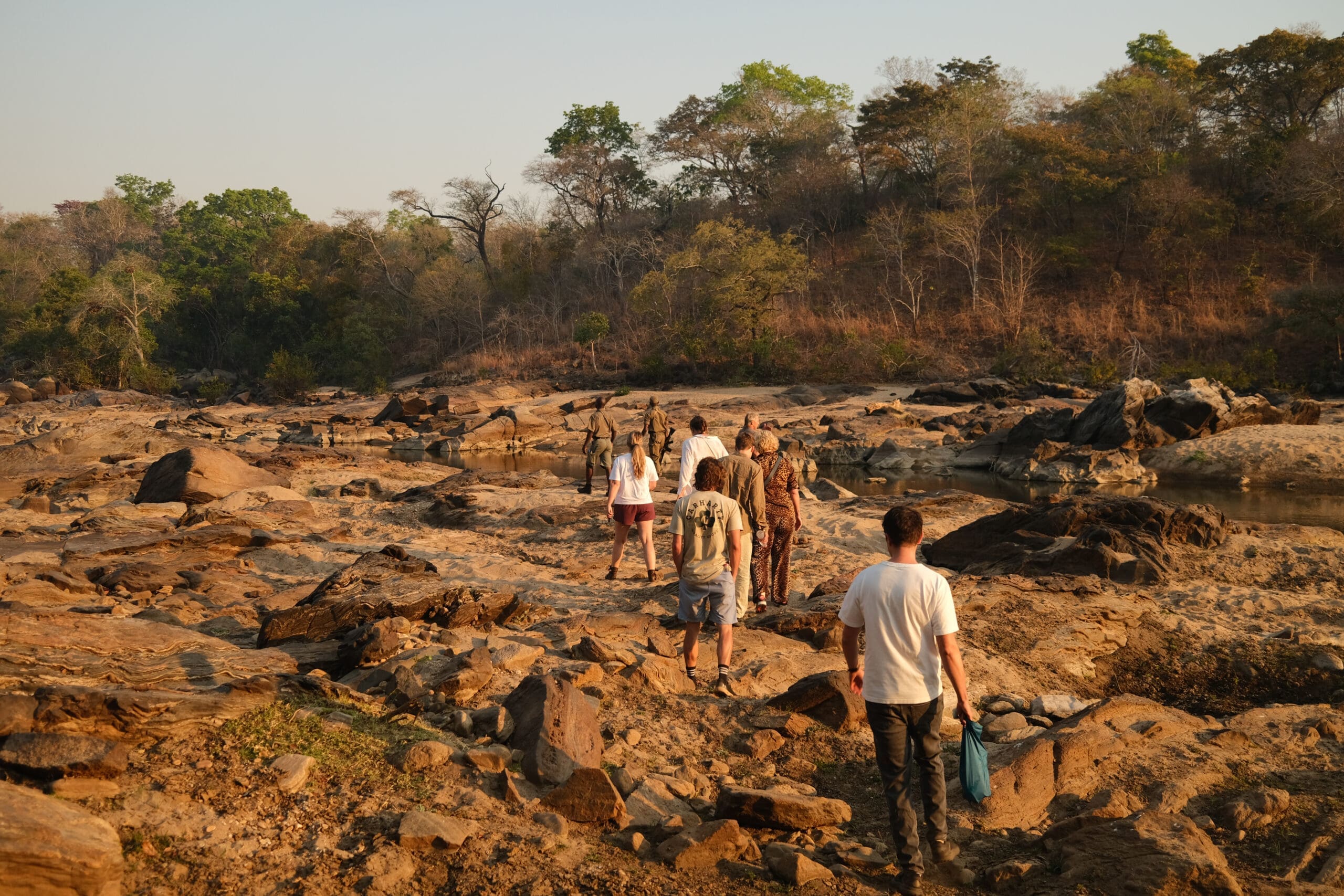 Een groep mensen, sommigen in safarikleding, loopt in één rij over een rotsachtige, droge rivierbedding in Malawi, omringd door schaarse bomen en struiken onder een heldere Afrikaanse hemel - perfect voor avontuurlijke reizen.