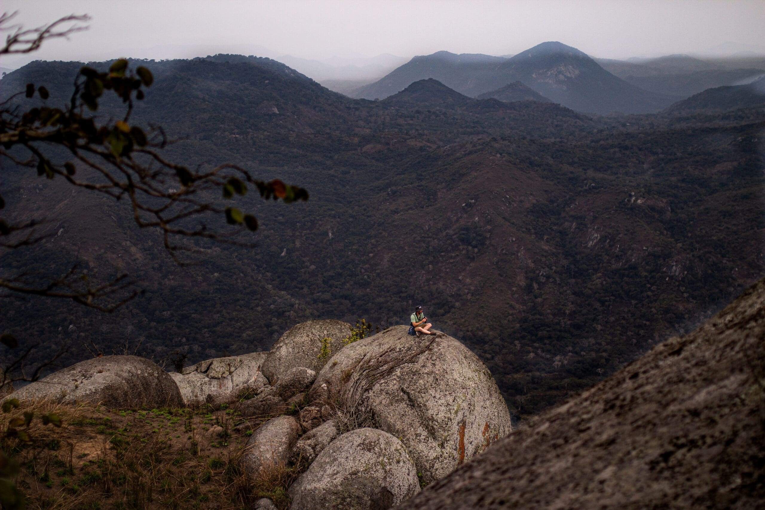 Een persoon zit alleen op een grote rotsformatie in Angola, omringd door ruige bergen en dichte bossen onder een bewolkte hemel. Dit creëert een gevoel van eenzaamheid in het uitgestrekte Afrikaanse landschap dat reizigers aantrekt.