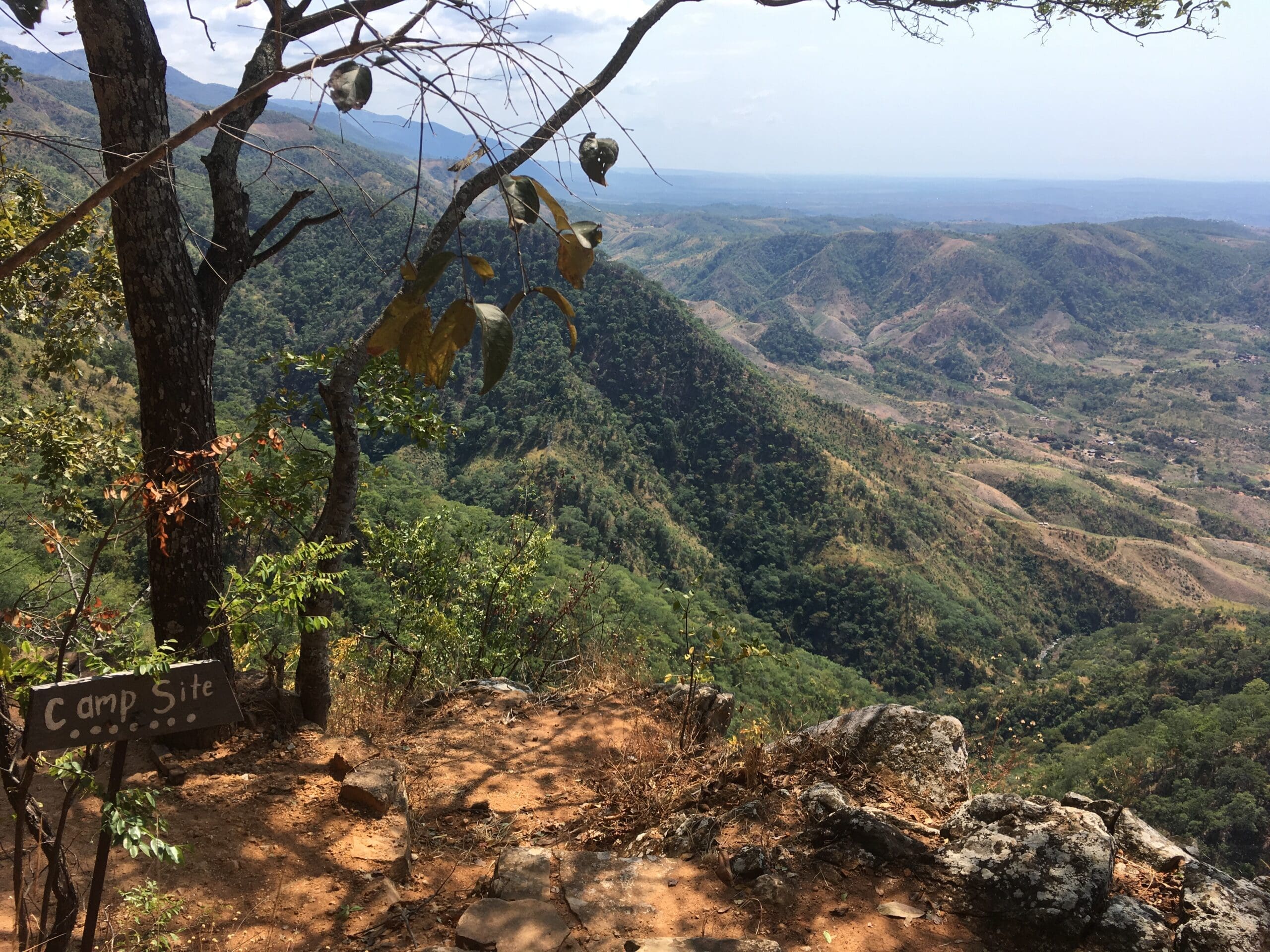 Een schilderachtig uitzicht op de bergen in Malawi met een zandpad dat naar een klif leidt, omringd door bomen en groen. Links aan een boom hangt een houten bord met de tekst Camp Site. Verken de valleien en heuvels van Afrika die zich in de verte uitstrekken - perfect om te reizen.