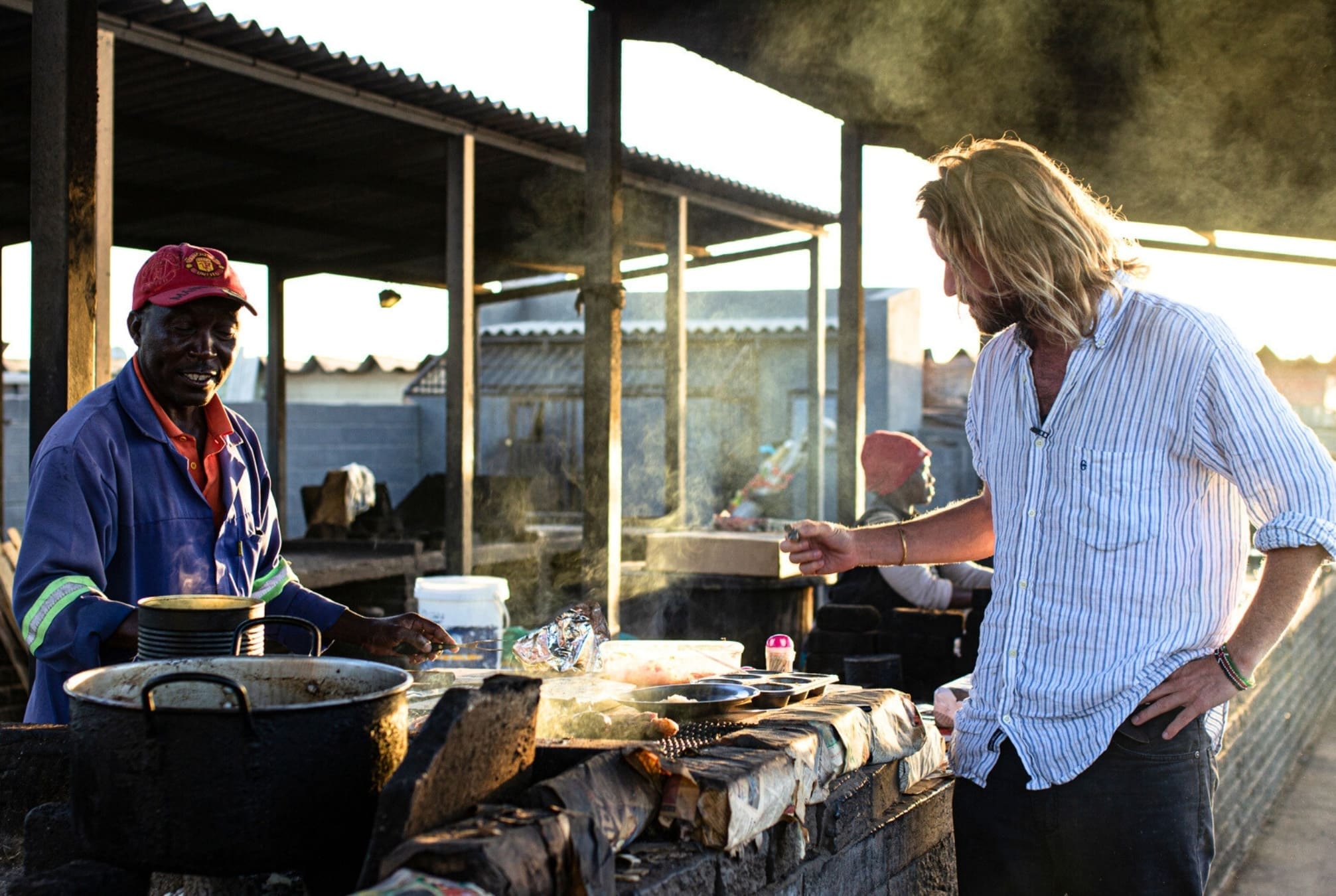 Ein Mann mit roter Mütze kocht an einem Imbissstand mit großen Töpfen und rauchigen Grills, während ein anderer Mann mit langen Haaren und einem gestreiften Hemd in der Nähe steht und sich unterhält. Das Sonnenlicht fällt von der Seite ein.