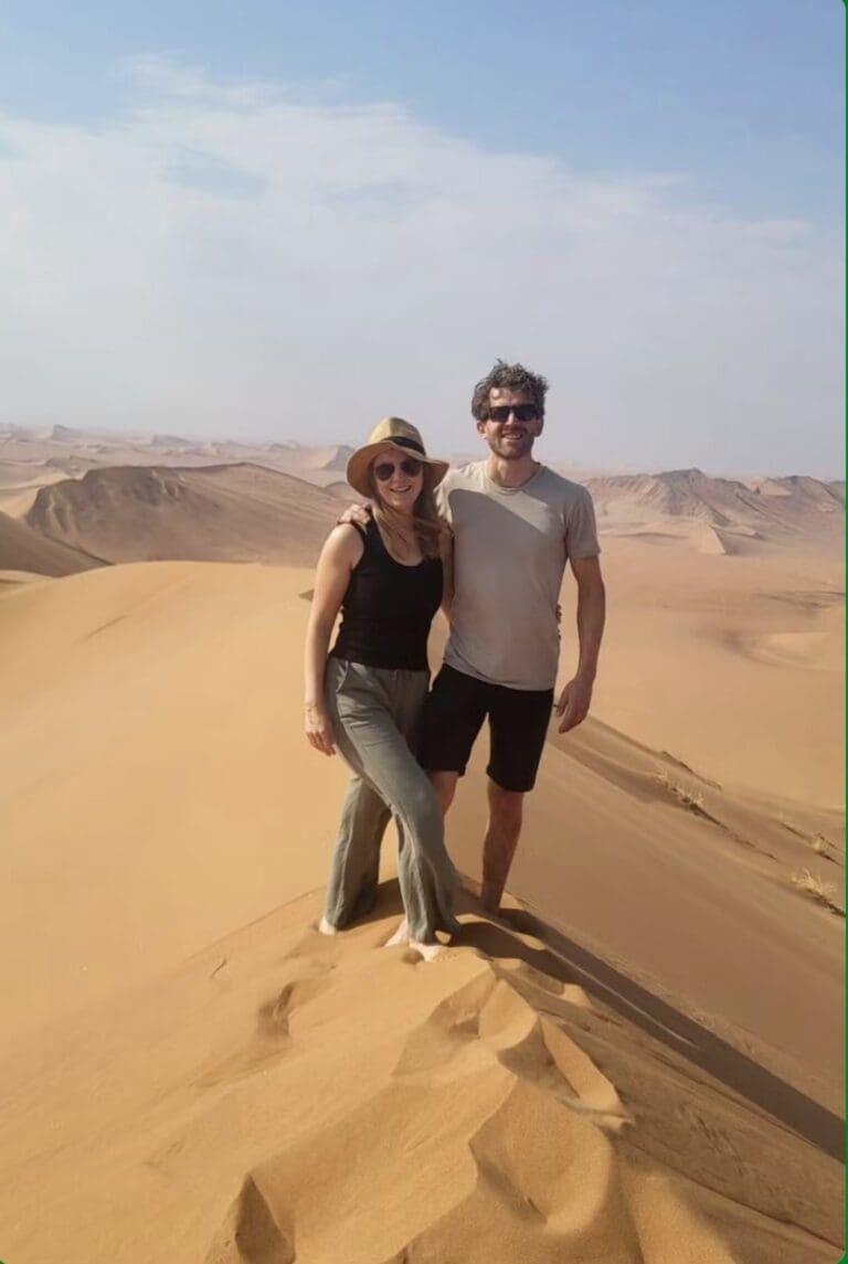 Eine Frau und ein Mann stehen zusammen auf einer großen Sanddüne und lächeln in die Kamera. Die Landschaft um sie herum besteht aus wogenden Sanddünen unter einem klaren Himmel.