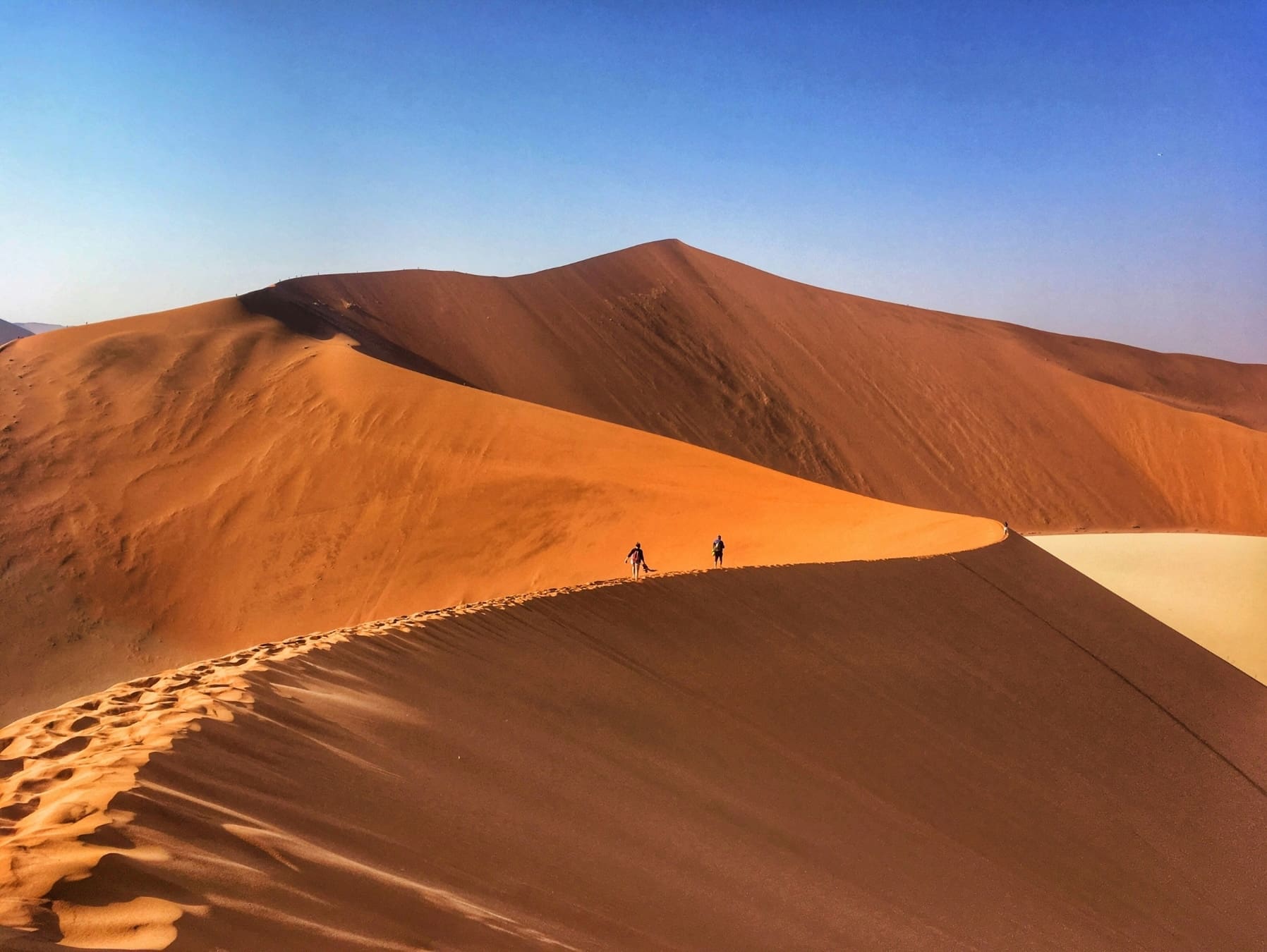 Zwei Personen spazieren auf dem Kamm einer großen Sanddüne unter blauem Himmel, umgeben von weiten, sonnenbeschienenen Wüstendünen.