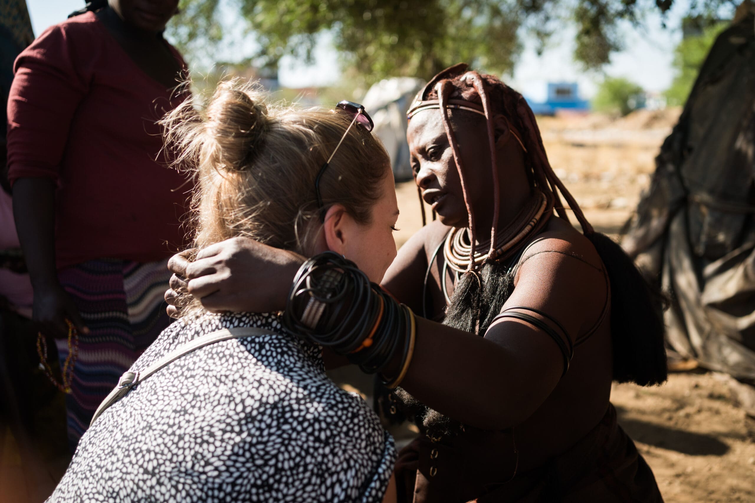 Een Himba-vrouw, getooid met traditionele sieraden en haarversieringen, houdt voorzichtig de schouders van een bezoeker vast terwijl ze in haar ogen kijkt, terwijl een andere persoon op de achtergrond in de buurt staat.