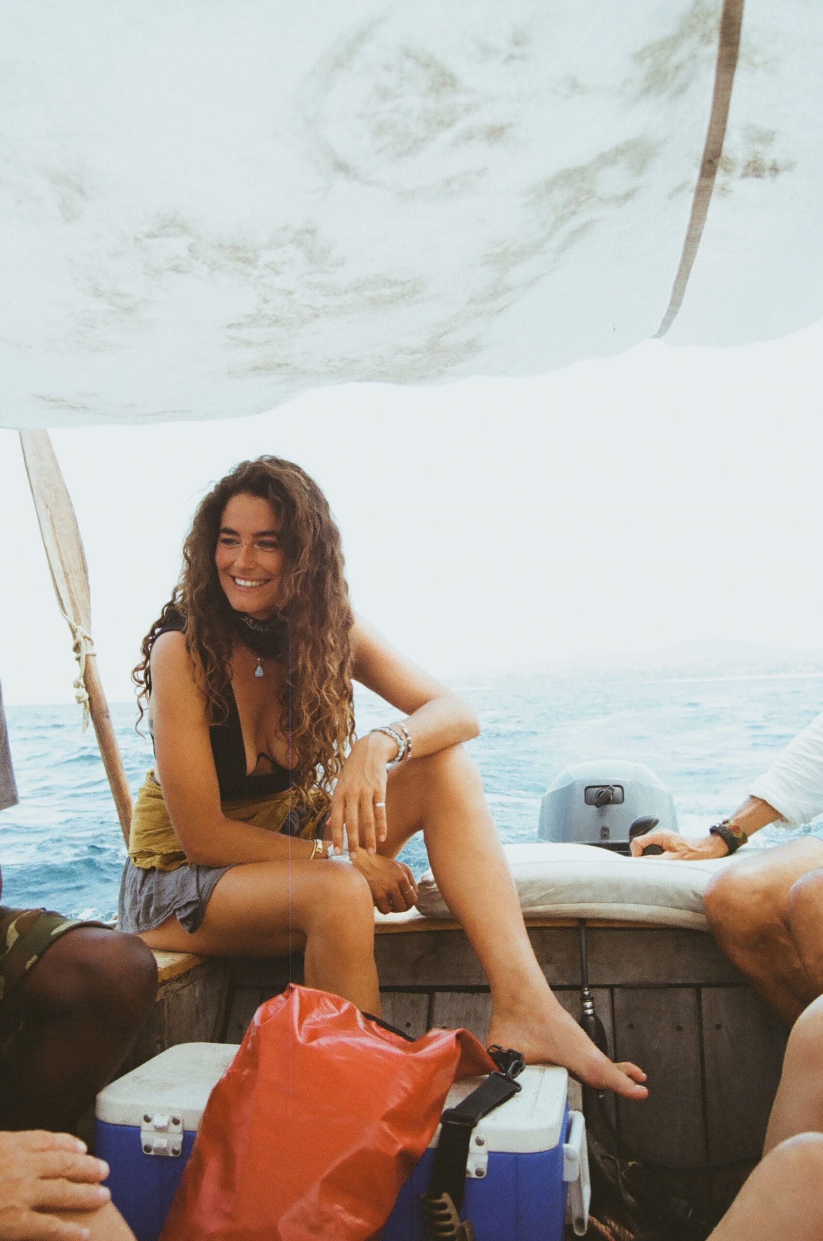Een vrouw met lang krullend haar zit glimlachend op een boot onder een afdak, omringd door tassen en andere mensen, met de oceaan zichtbaar op de achtergrond.