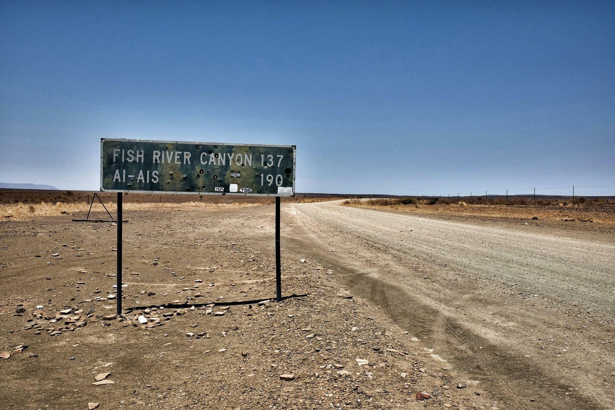Een verweerd verkeersbord in een droog, kaal landschap wijst naar Fish River Canyon (137 km) en Ai-Ais (190 km). Een onverharde weg strekt zich in de verte uit onder een strakblauwe hemel.