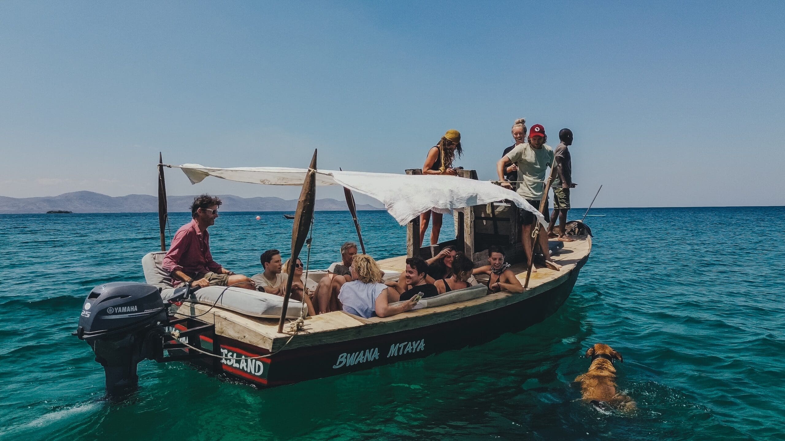 Een groep mensen ontspant zich op een kleine houten boot met luifel, drijvend op helderblauw water. Een hond zwemt vlakbij. De lucht is helder en zonnig.