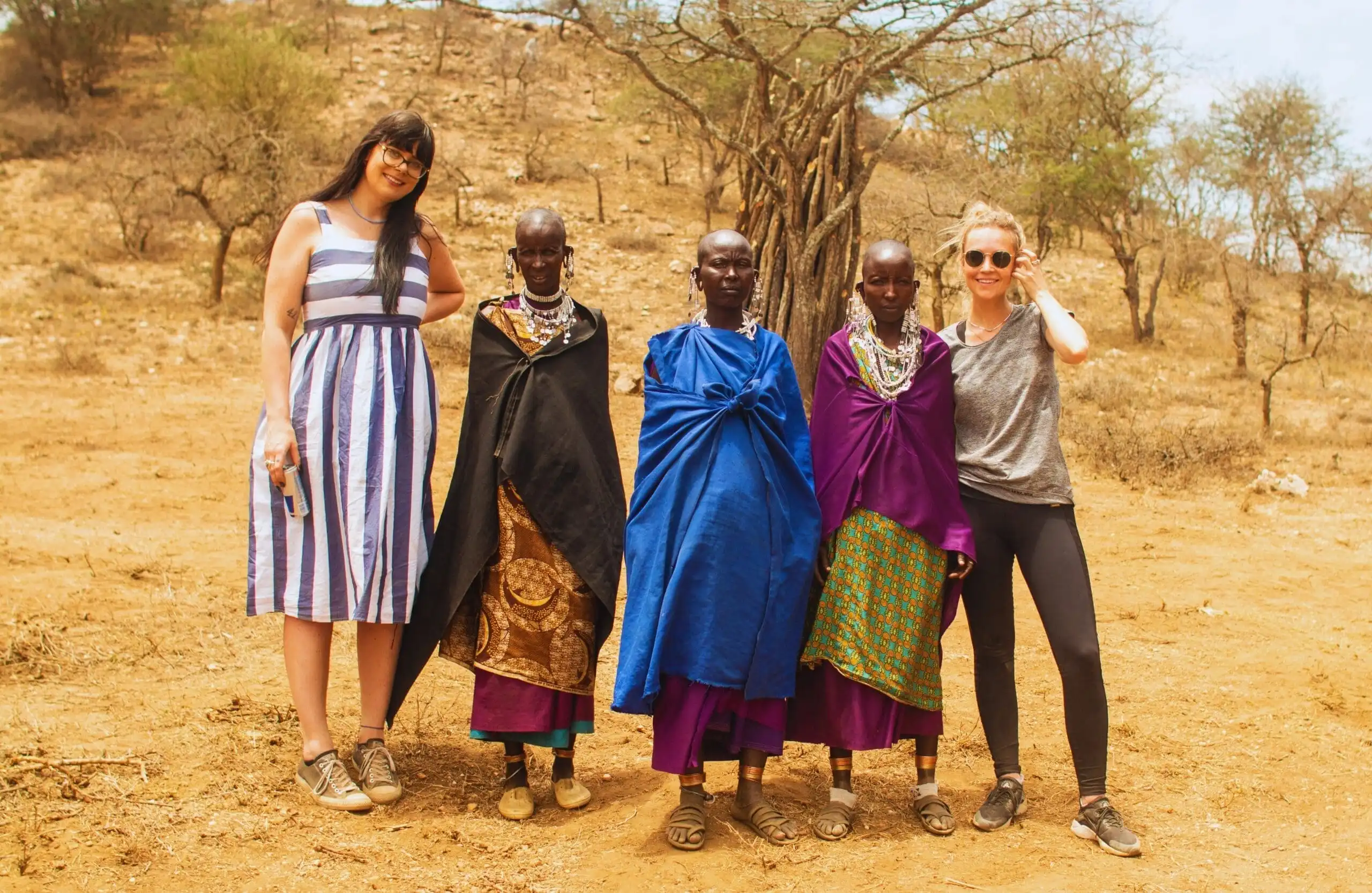Twee vrouwen in moderne kleding staan naast drie Maasai-vrouwen in traditionele kleurrijke kleding en sieraden, poserend voor een foto in een droog, landelijk landschap met schaarse bomen.