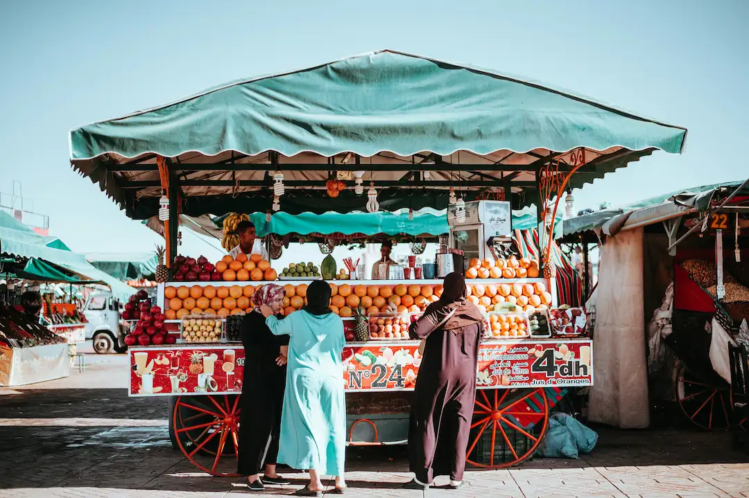 Een kleurrijk fruitstalletje met een groen afdak op een openluchtmarkt. Vooraan staan drie vrouwen in traditionele kleding fruit uit te zoeken. Verse sinaasappels, citroenen en ander fruit liggen netjes opgestapeld op de kar.