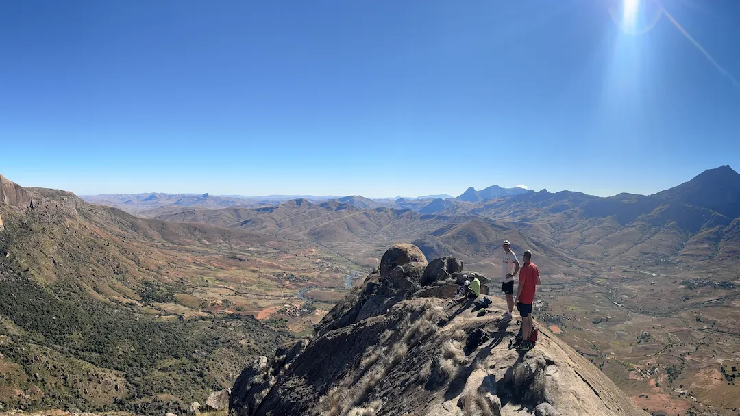 Drie wandelaars staan op de top van een rotsachtige berg en kijken uit over een uitgestrekte, zonovergoten vallei met glooiende heuvels en bergen in de verte, onder een helderblauwe hemel. Eén van hen zwaait naar de camera.