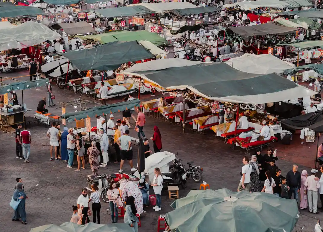 Een scène op een openluchtmarkt met veel kraampjes die met groene zeilen zijn afgedekt, verkopers die hun waren aanbieden en menigten mensen die rondlopen, winkelen en met elkaar praten. Op sommige tafels liggen kleurrijke spullen uitgestald en tussen de winkelende mensen staan motorfietsen geparkeerd.