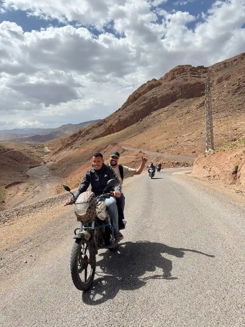 Twee mensen rijden op een motorfiets over een bochtige, verlaten weg door een rotsachtig, bergachtig landschap onder een gedeeltelijk bewolkte hemel. In de verte volgen nog meer motorrijders.
