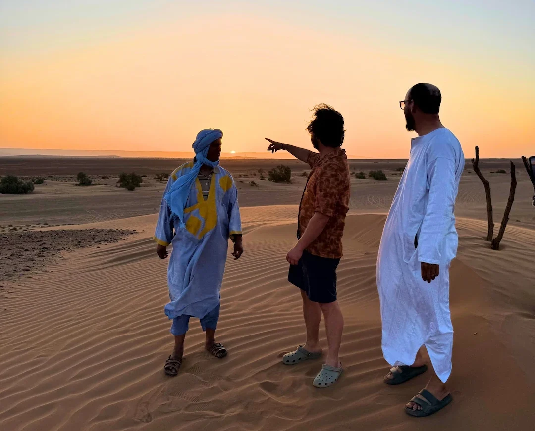 Drie mannen staan bij zonsondergang op het woestijnzand; één van hen wijst naar de horizon. Ze dragen verschillende soorten traditionele en moderne kleding, met op de achtergrond duinen, schaarse begroeiing en een gloeiende hemel.