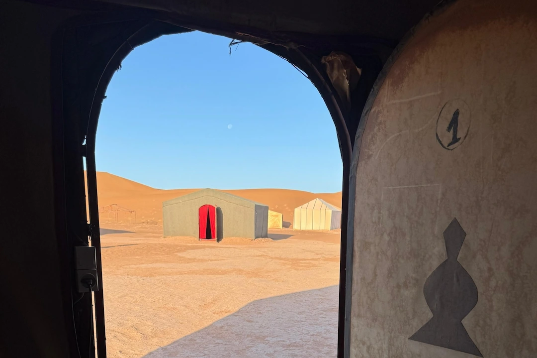 Uitzicht vanuit een tent op andere tenten in een woestijn met een zandgrond en duinen op de achtergrond, onder een helderblauwe hemel.