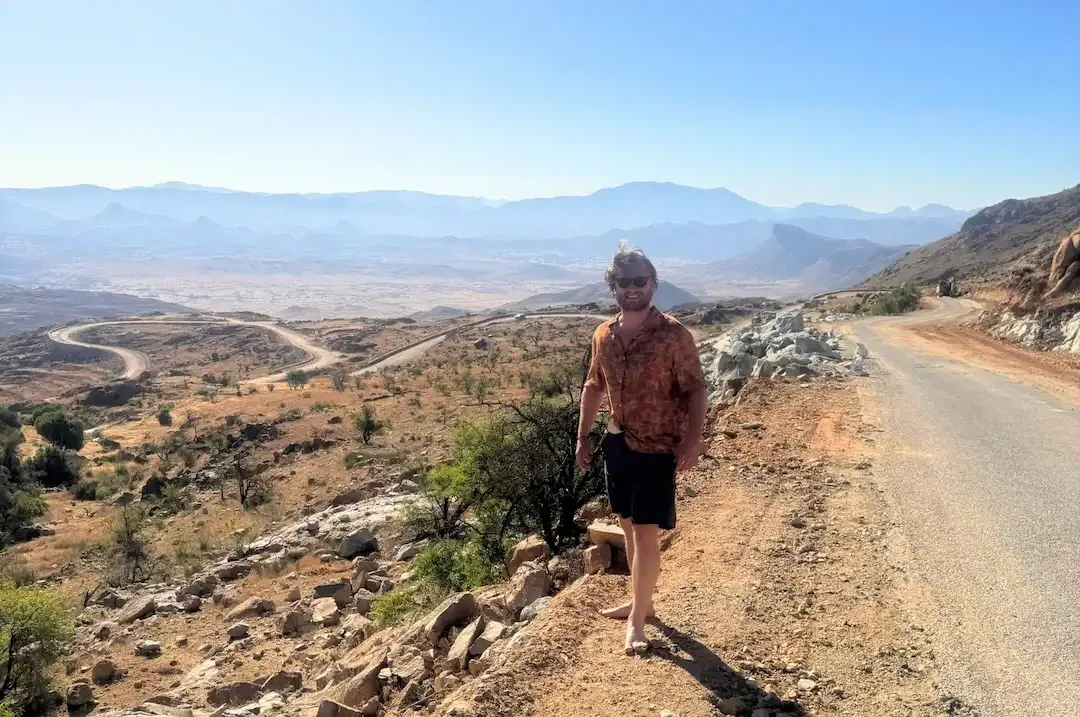 Een man in een shirt met patroon en een korte broek staat op blote voeten naast een landweg, uitkijkend over een droog, rotsachtig landschap met kronkelende wegen en bergen in de verte onder een helderblauwe hemel.