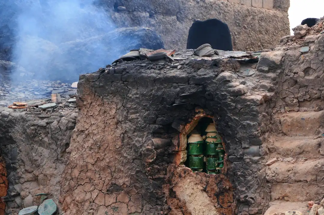 In een traditionele oven met een ruwe klei-buitenkant wordt ongebakken aardewerk gebakken, terwijl op de achtergrond rook opstijgt, wat duidt op het bakproces.