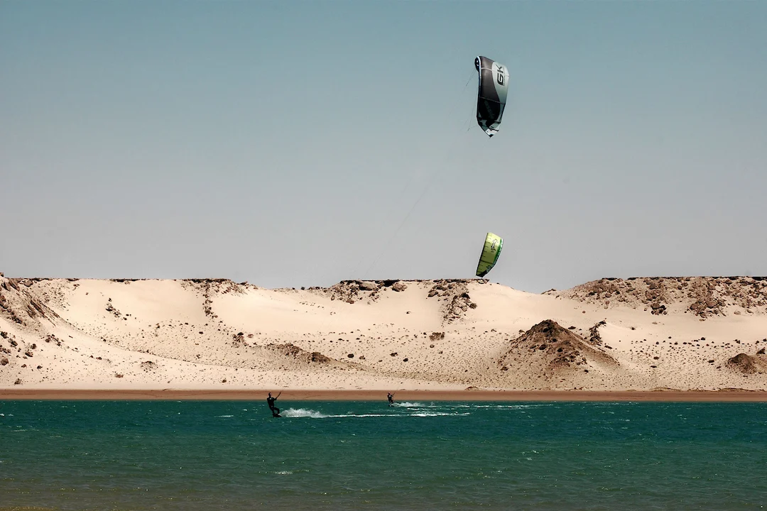 Twee mensen zijn aan het kitesurfen op het turkooizen water bij de zandduinen, onder een strakblauwe hemel.