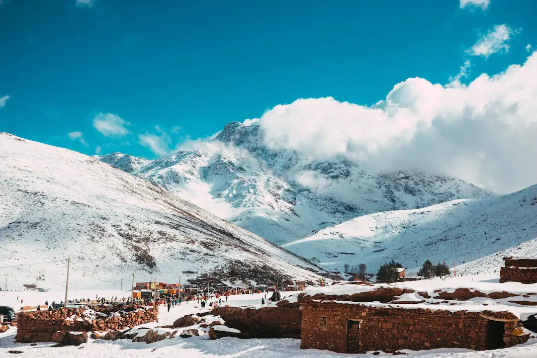Met sneeuw bedekte bergen onder een stralend blauwe lucht met hier en daar wat wolken; mensen die zich verzamelen bij rustieke stenen gebouwen in de besneeuwde vallei beneden.