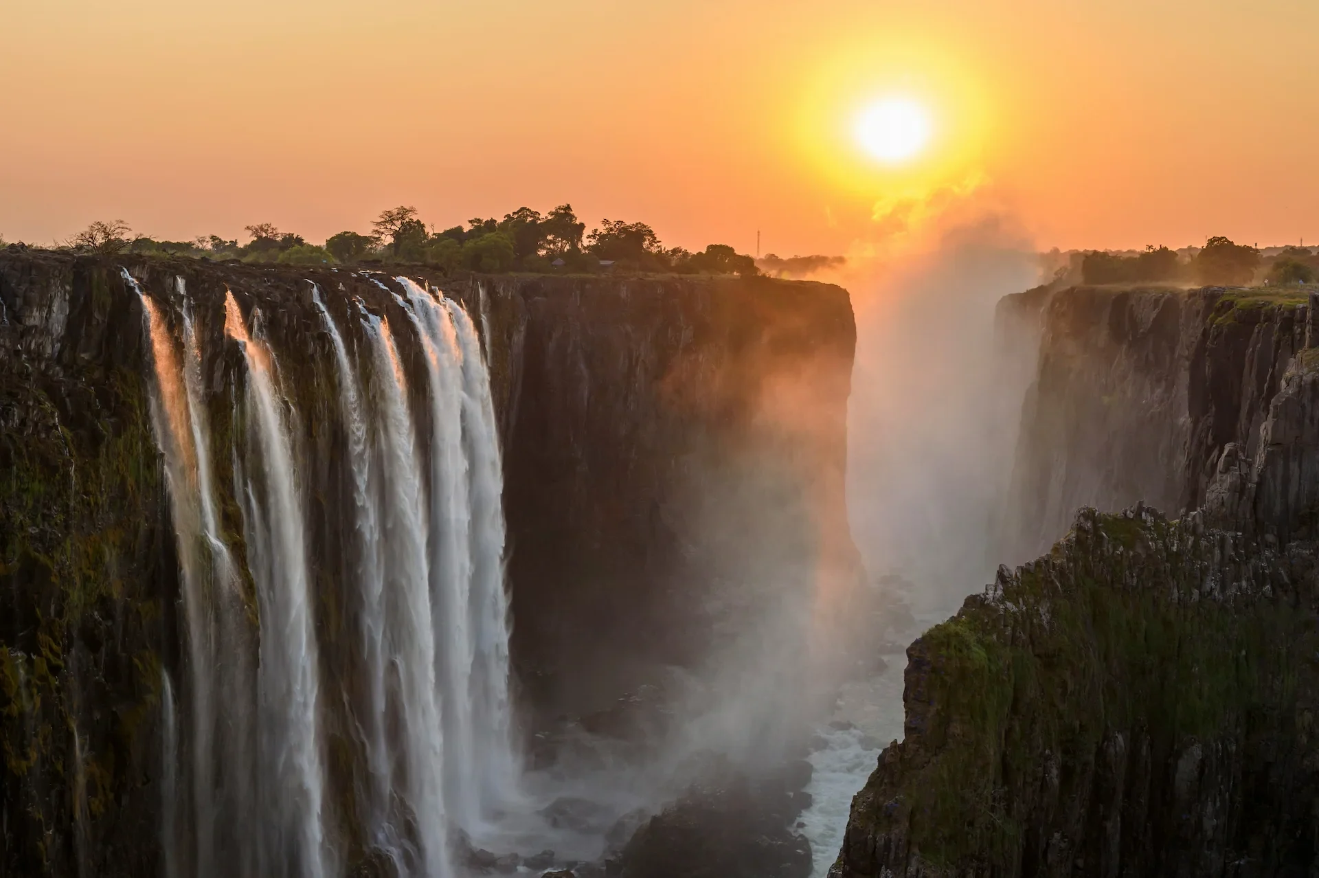 Het water stort zich via een brede waterval naar beneden in een diepe kloof, terwijl er nevel opstijgt en de zon op de achtergrond ondergaat, waardoor het landschap in een warm, goudkleurig licht wordt gehuld.
