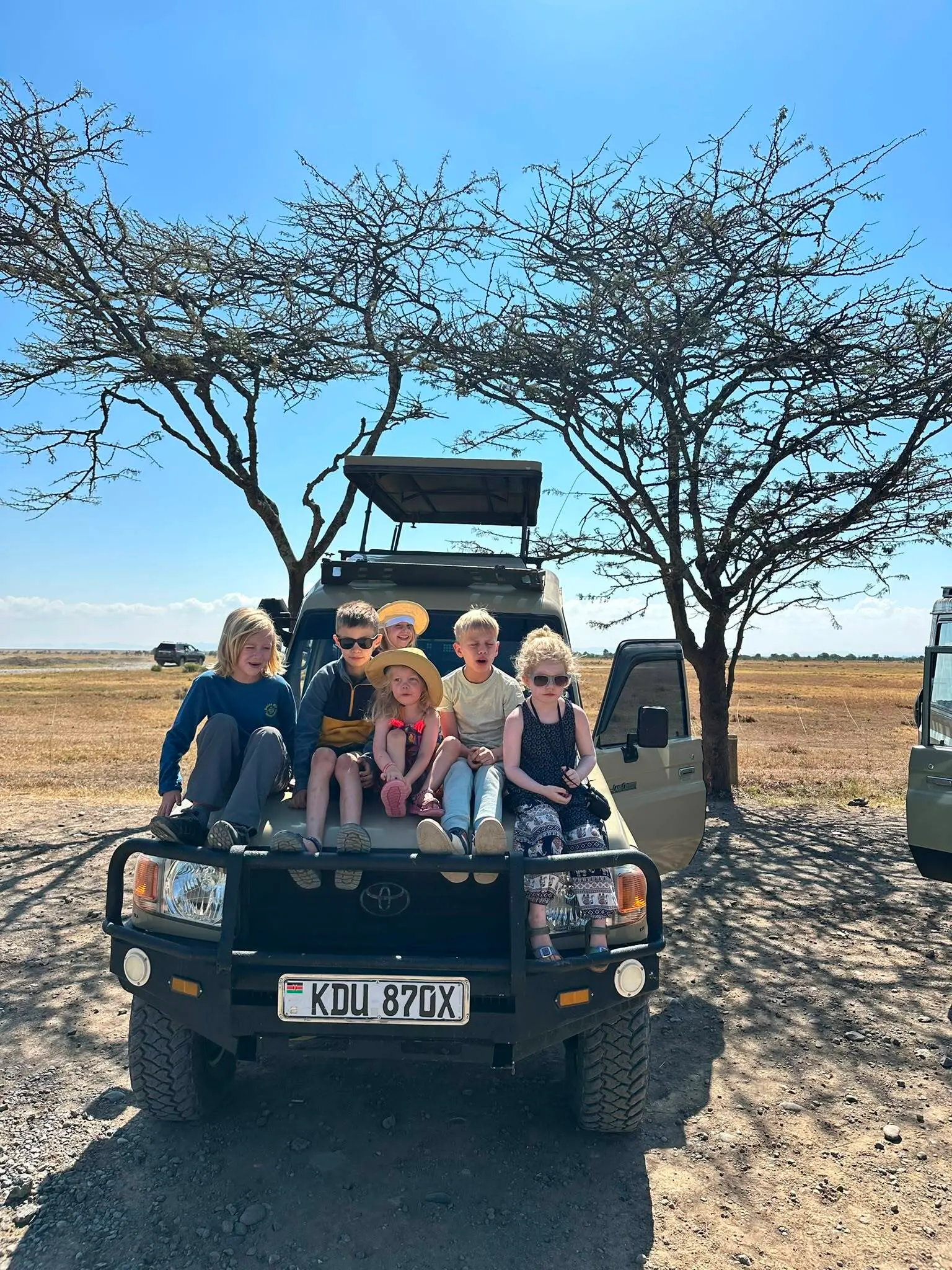 Zes kinderen zitten voorin een safarijeep, die op een droge vlakte onder twee kale bomen geparkeerd staat. De lucht is helder en blauw, en de kinderen dragen hoeden, zonnebrillen en vrijetijdskleding.