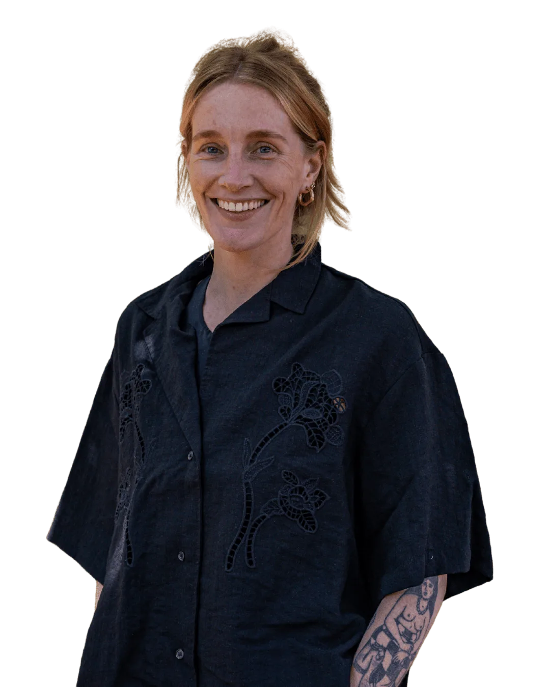 Een persoon met licht haar, gekleed in een donker shirt met korte mouwen en bloemenborduursels, lacht naar de camera. Op de linkeronderarm zijn tatoeages te zien. De achtergrond is effen wit.