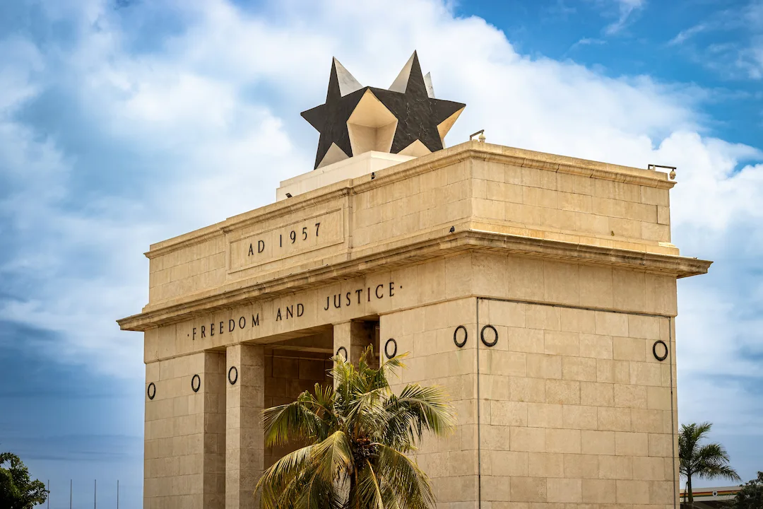 Een close-up van de Onafhankelijkheidsboog van Ghana, met daarbovenop een zwarte ster. Op het opschrift staan de jaartal 1957 en de woorden ‘Vrijheid’ en ‘Rechtvaardigheid’. Op de achtergrond zijn palmbomen en een gedeeltelijk bewolkte hemel te zien.