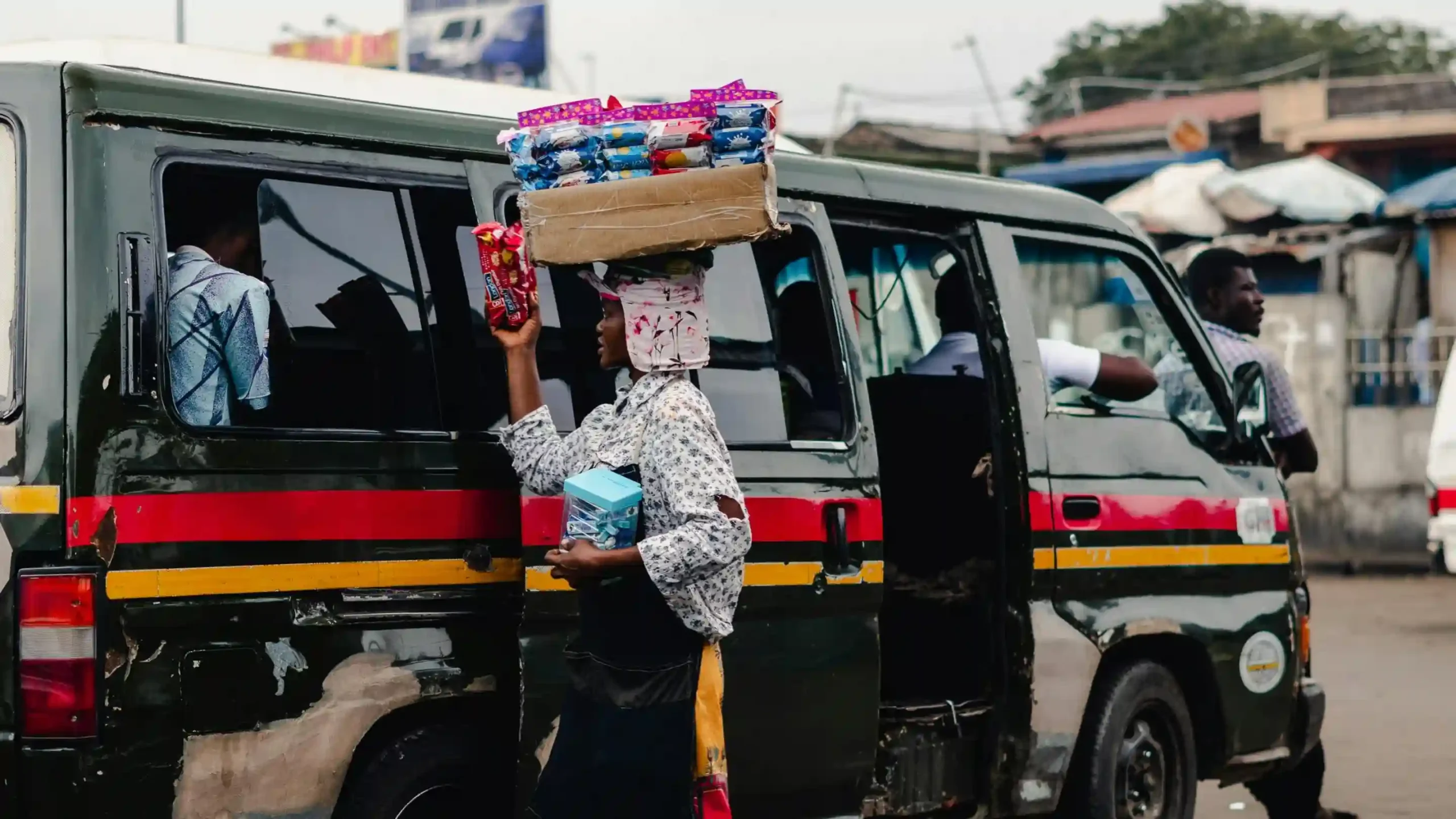 Een straatverkoopster balanceert snacks op haar hoofd en verkoopt spulletjes aan passagiers door het raam van een minibus. De deuren van de minibus staan open en je kunt de mensen binnen zien zitten. Op de achtergrond zijn stadsgebouwen te zien.