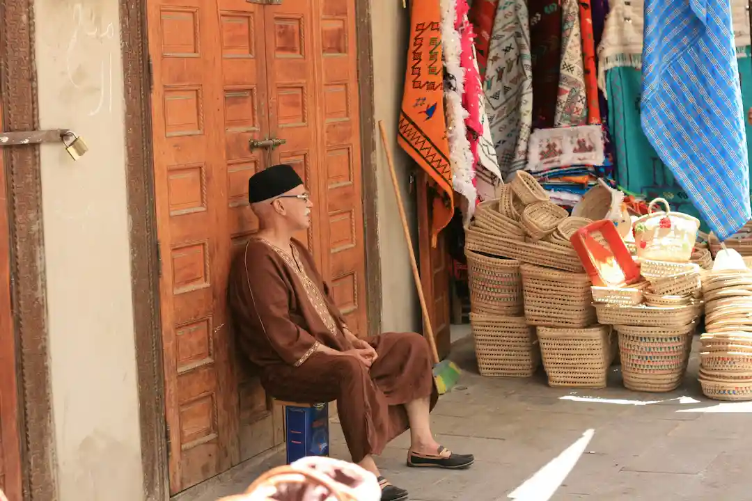 Een oudere man in bruine traditionele kleding en met een zwarte pet zit op een kist naast een houten deur. Vlakbij liggen gevlochten manden en kleurrijke stoffen te koop uitgestald. Het zonlicht verlicht het markttafereel.