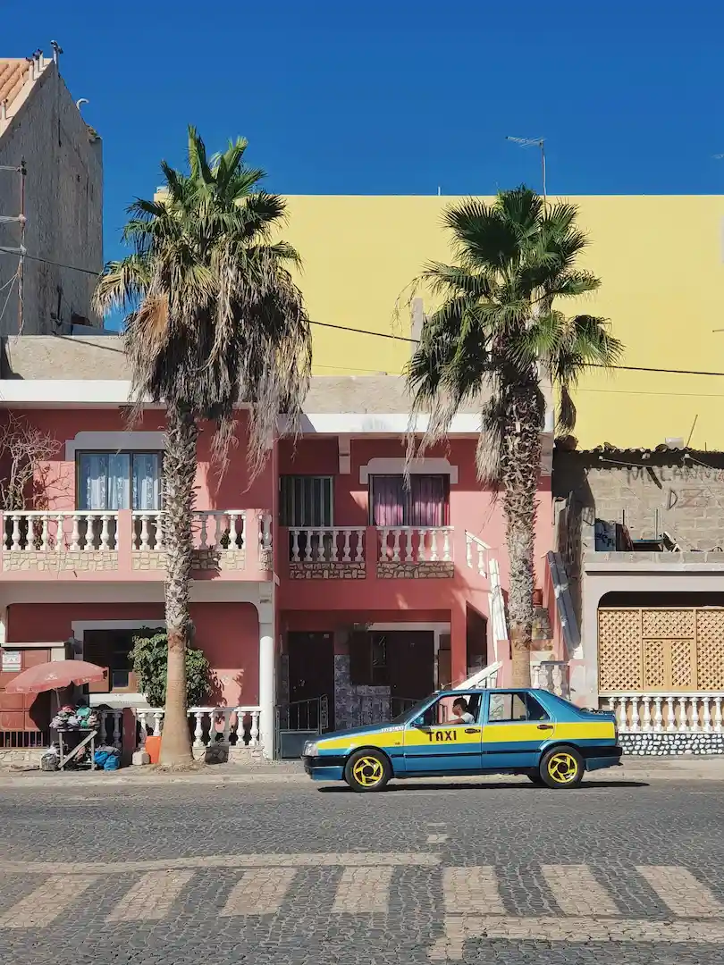 Een blauw-gele taxi staat geparkeerd op een straat met kasseien, voor een roze gebouw van twee verdiepingen met twee palmbomen en balkons, onder een helderblauwe hemel.